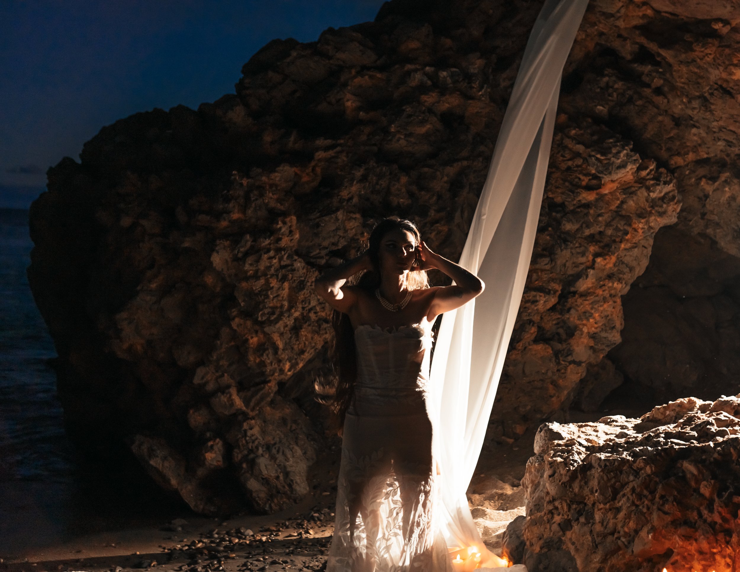 Une femme portant une robe blanche se tient sur une plage rocheuse la nuit, avec un tissu léger suspendu dans l'air, éclairée par une lumière douce, créant une ambiance mystérieuse et élégante.