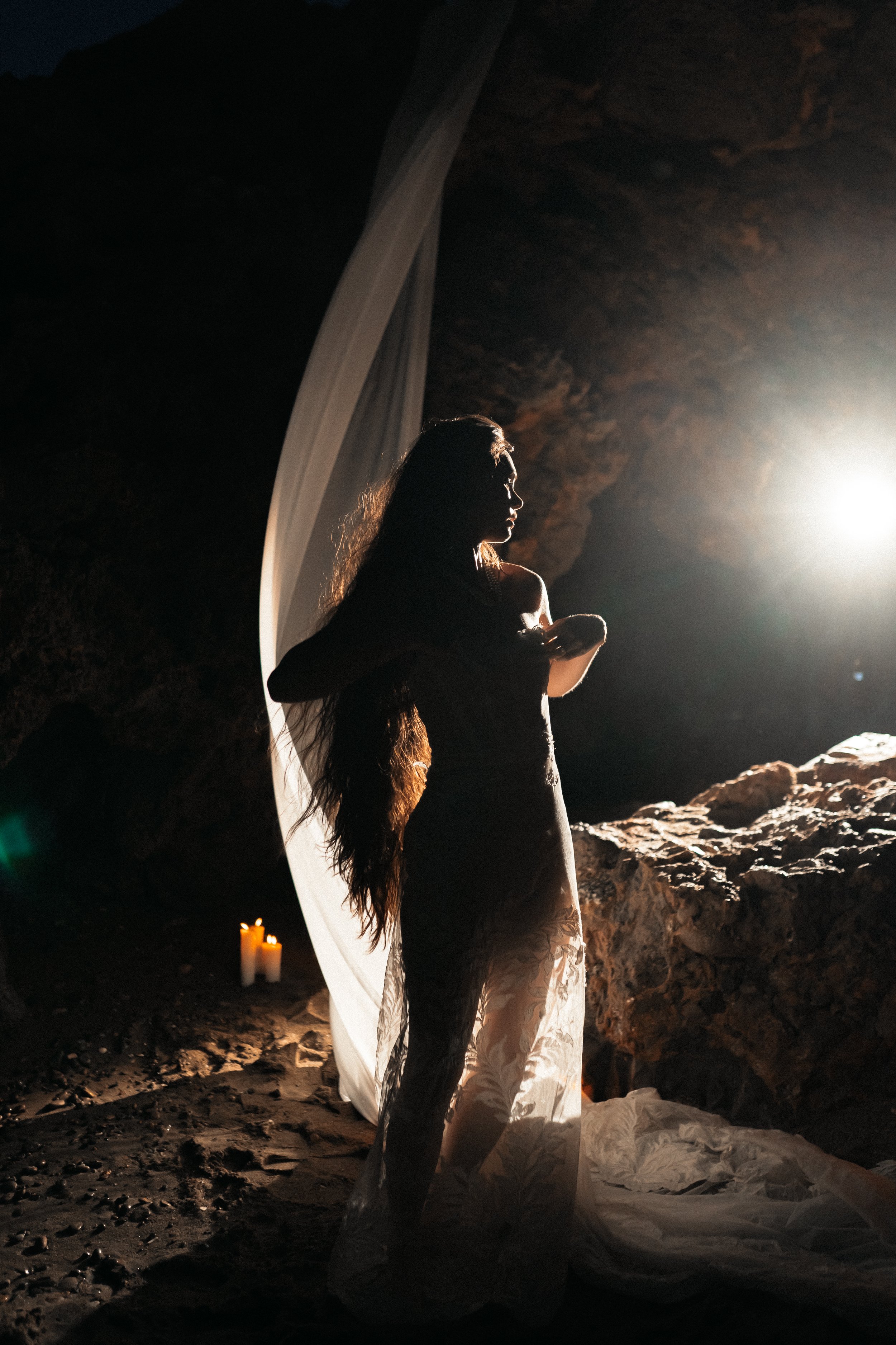 Silhouette d'une femme en robe longue dans une grotte, éclairée par une lumière intense à l'arrière-plan, avec des bougies allumées à côté.