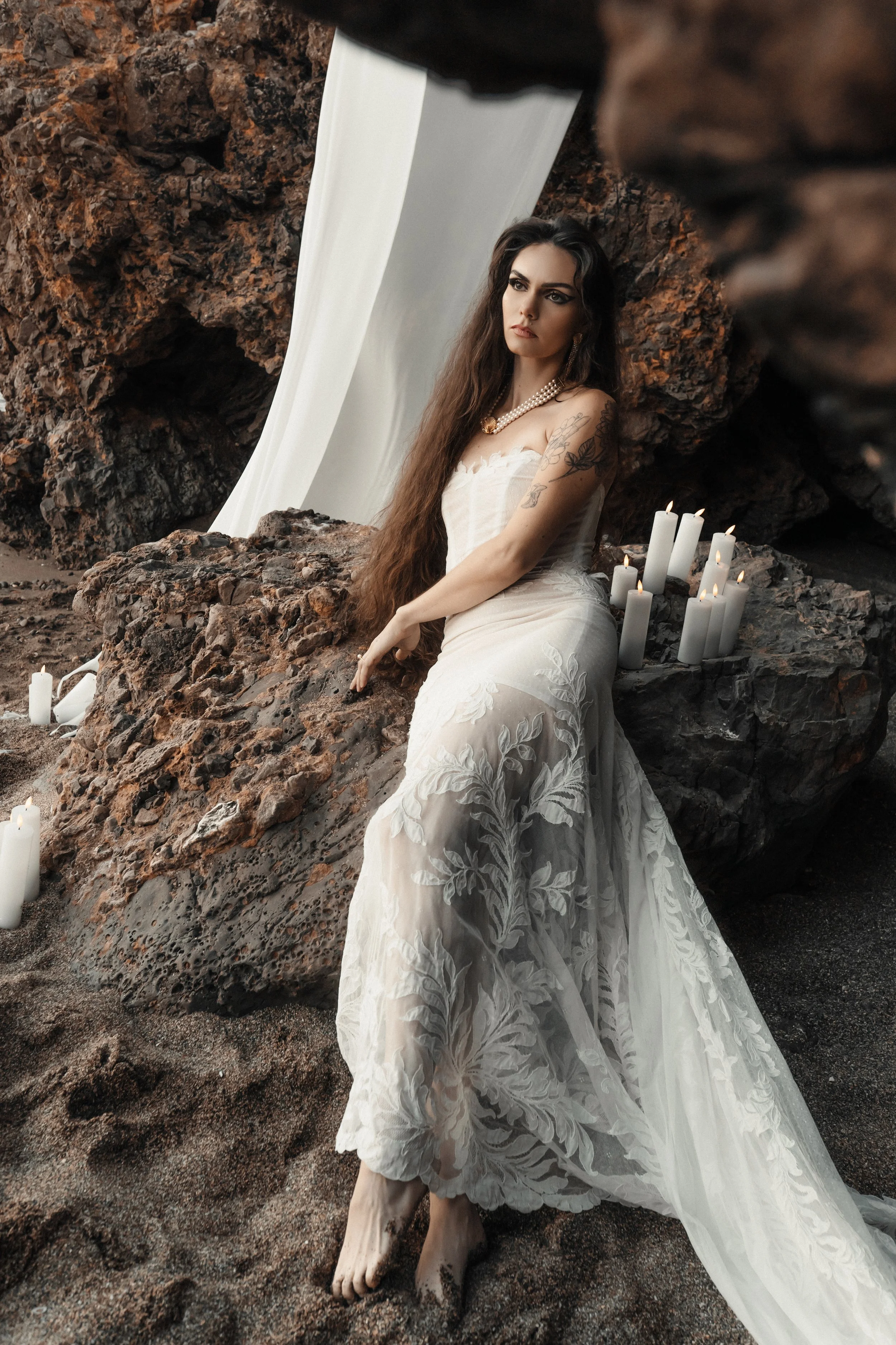 Une femme portant une robe de mariage blanche assise sur des rochers, entourée de bougies allumées, dans un environnement naturel avec des rochers noirs et un fond blanc.