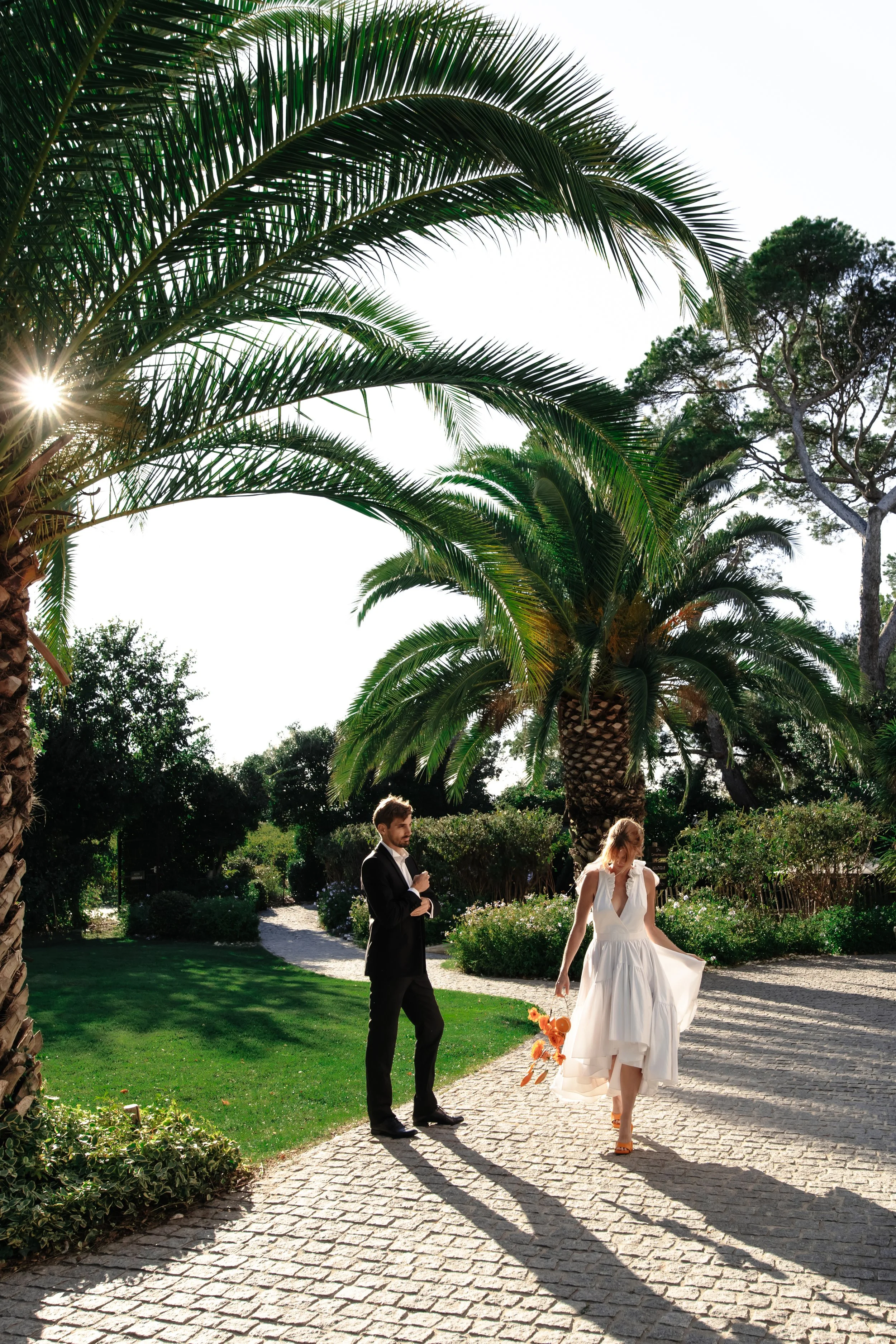 Un couple en tenue de mariage dans un petit jardin avec des palmiers et des arbres, soleil brillant.