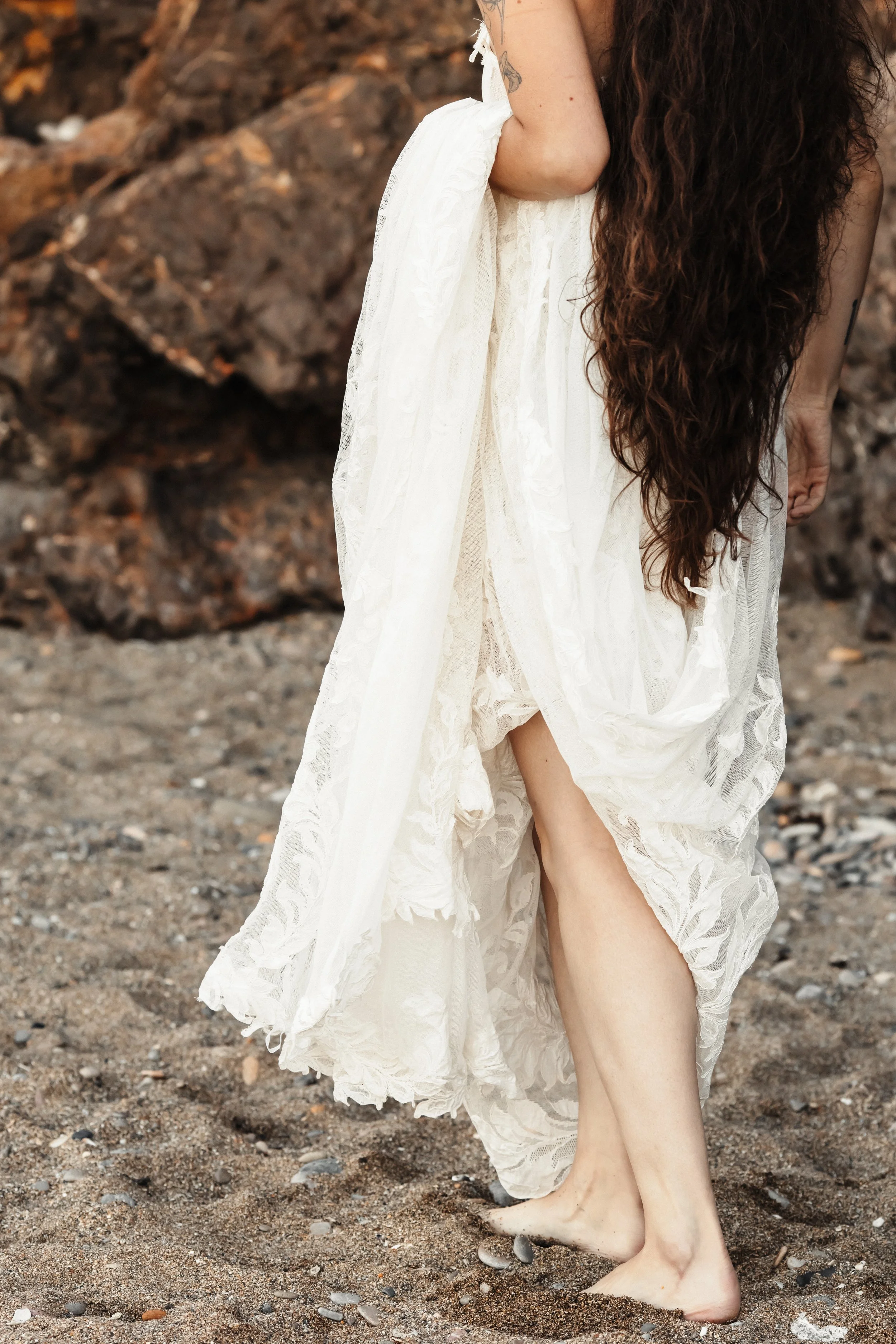 Femme portant une robe blanche en lace au sol, à la plage, avec des rochers en arrière-plan.