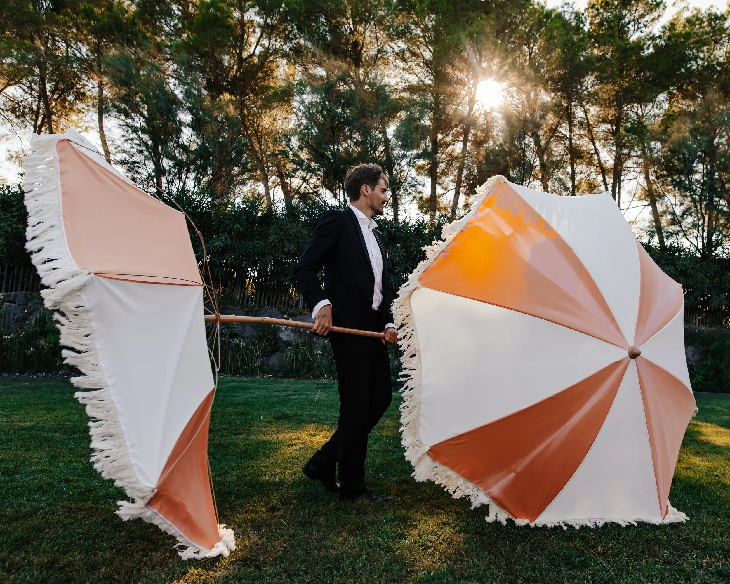 Un homme en costume tient deux parapluies géants à rayures orange et blanc dans un jardin avec des arbres en arrière-plan, sous un soleil brillant.