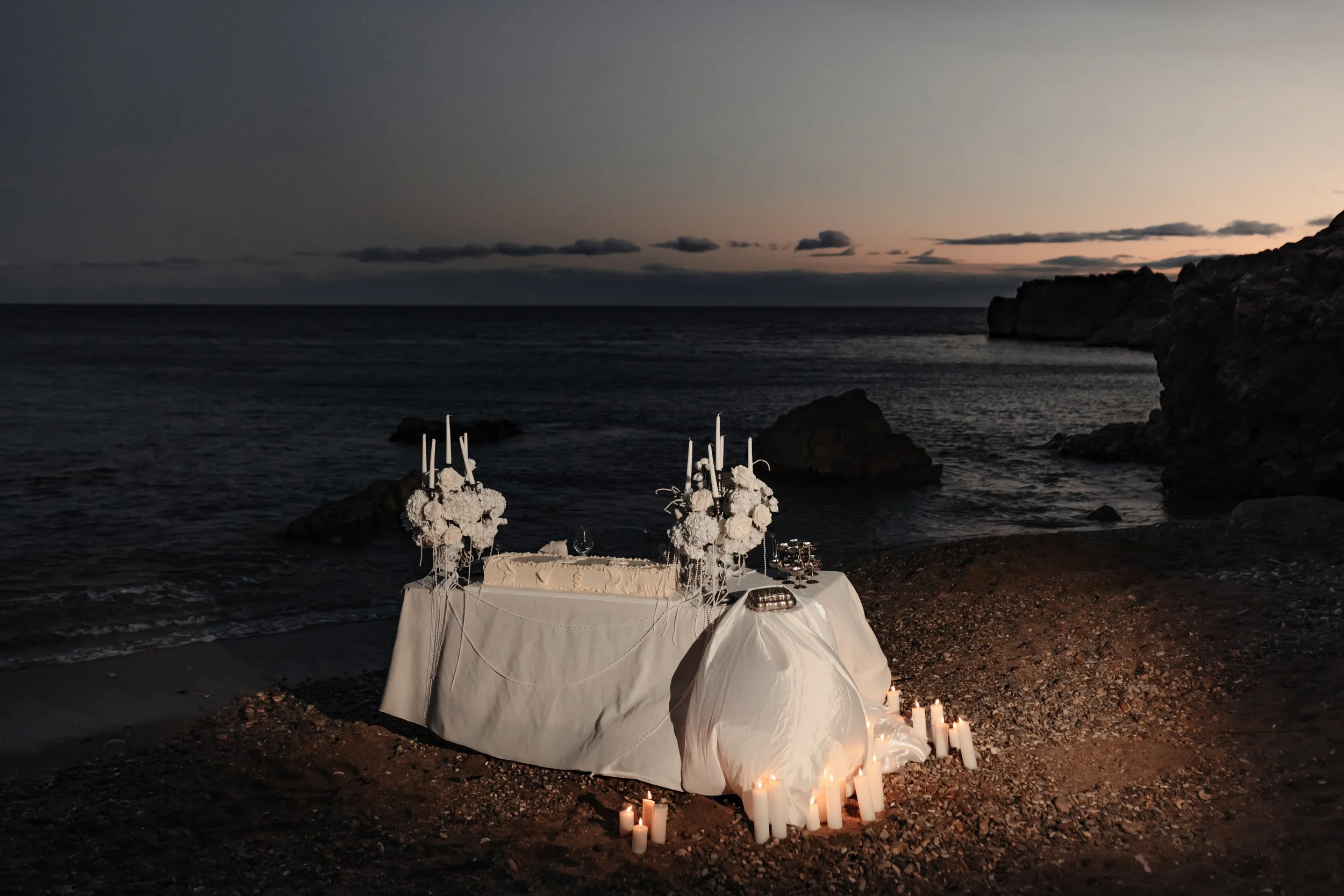 Un tableau décoré pour un événement ou un mariage, situé sur une plage lors du crépuscule, avec des bougies et des fleurs blanches.