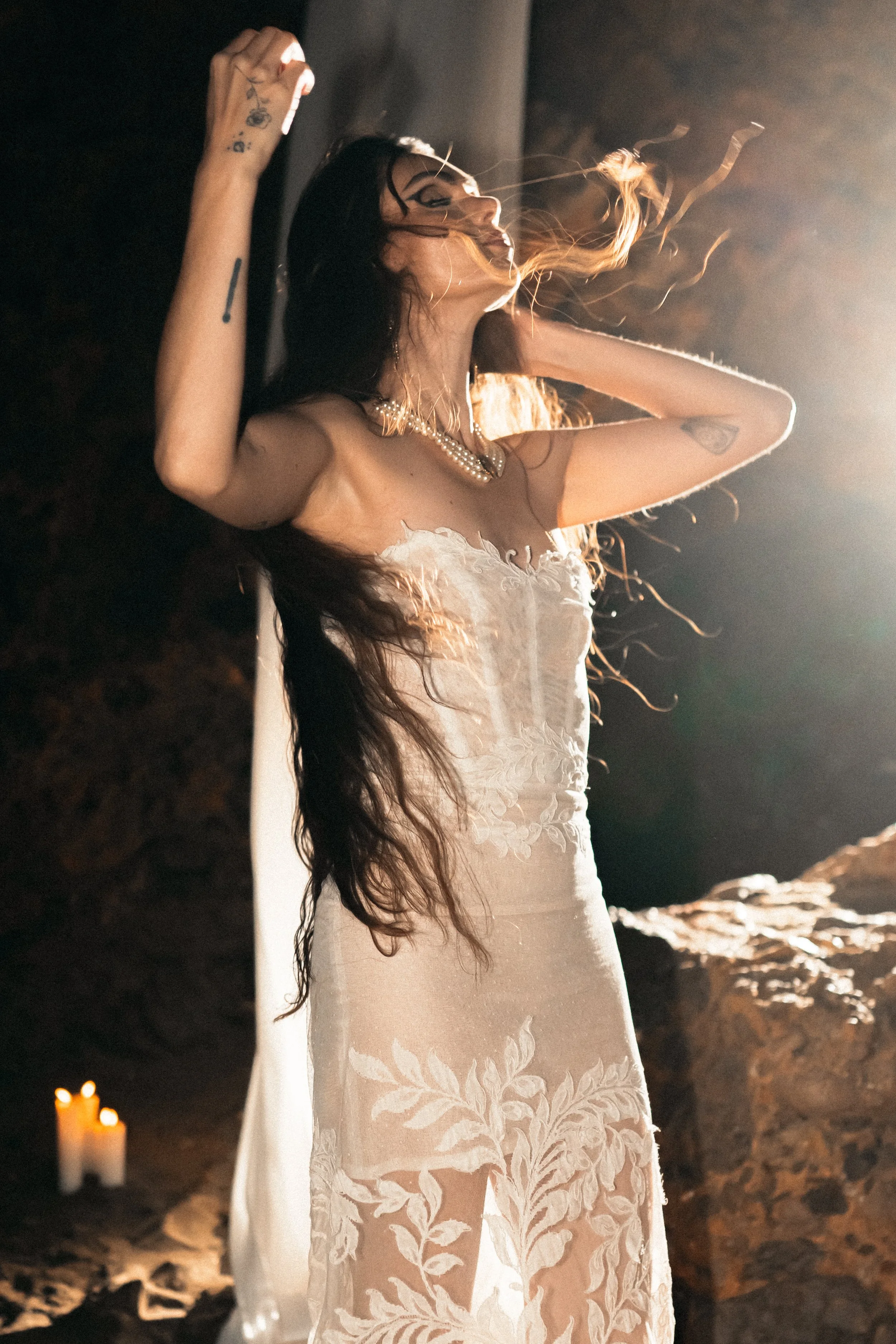 Femme portant une robe de mariée élégante, avec des cheveux longs et bruns, se tenant dans un endroit sombre avec de la lumière brillante derrière elle, et des bougies allumées en arrière-plan.