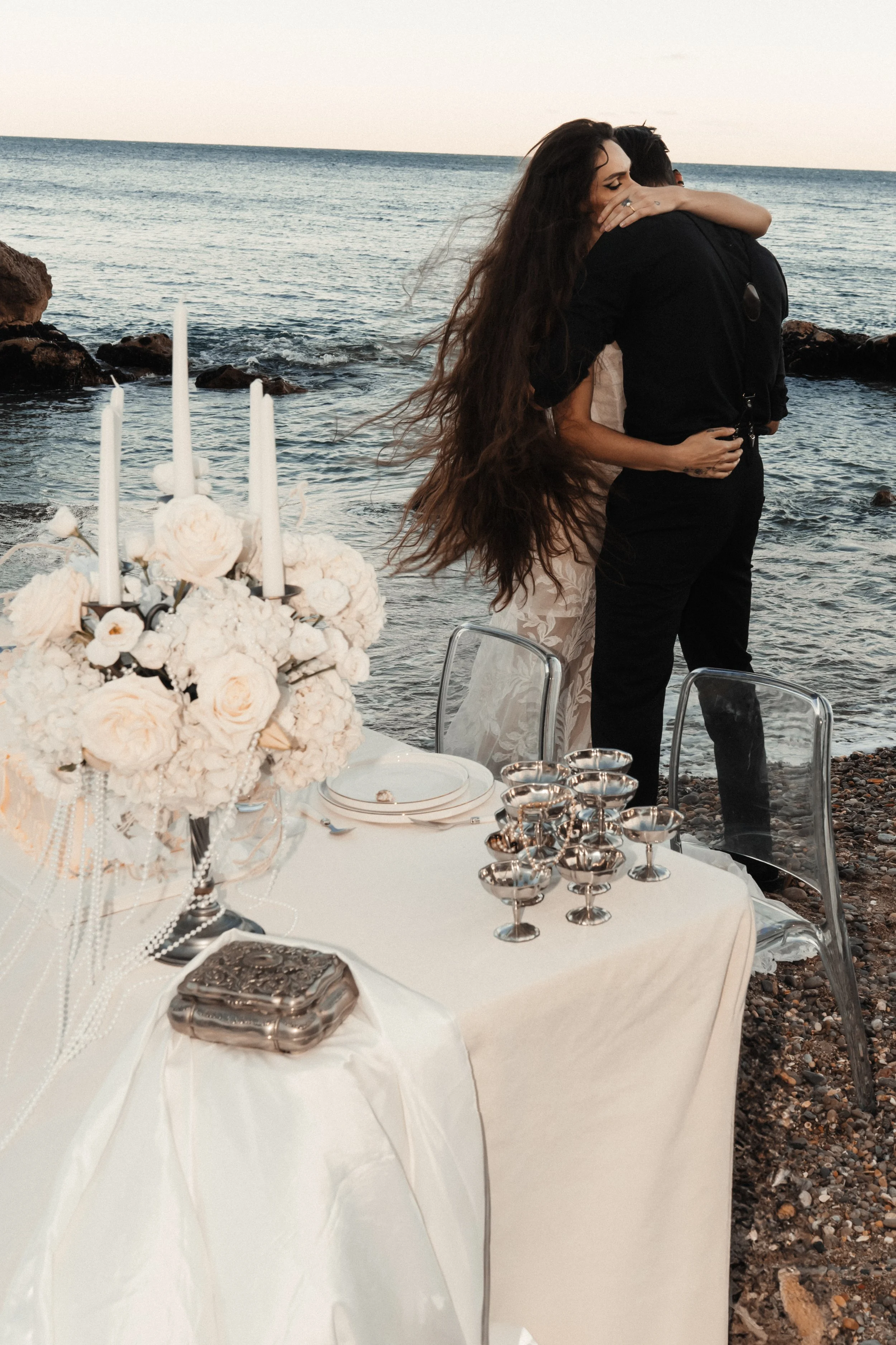 Un couple s'embrasse lors d'une cérémonie en plein air sur la plage, devant une table élégante ornée de fleurs blanches et de bougies, avec l'océan en arrière-plan.