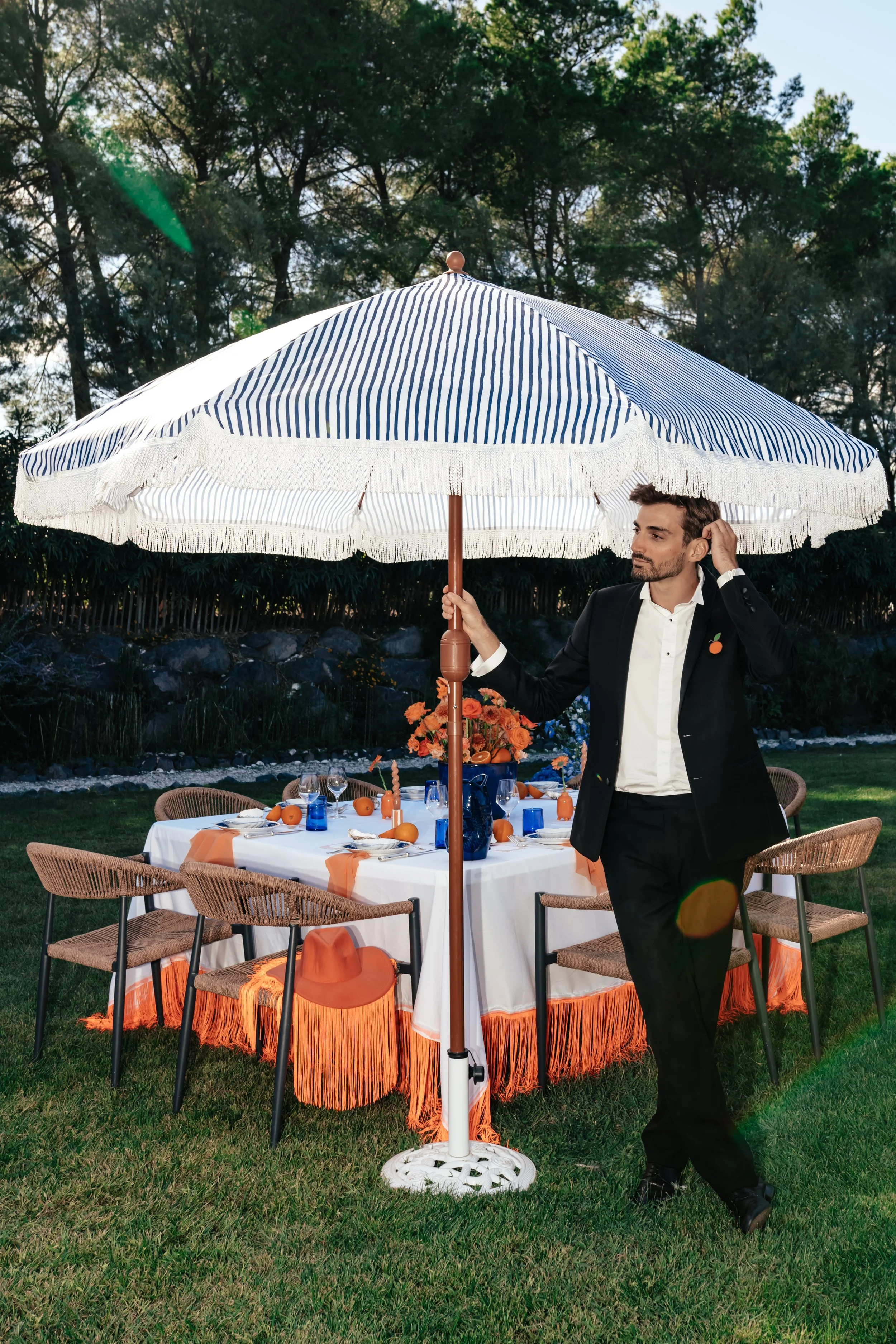 Un homme en costume noir se tient sous un grand parasol rayé blanc et bleu lors d'une fête en plein air. La table dressée est décorée de fleurs orange, bougies orange, assiettes, verres et serviettes orange, avec un hors tout orange.