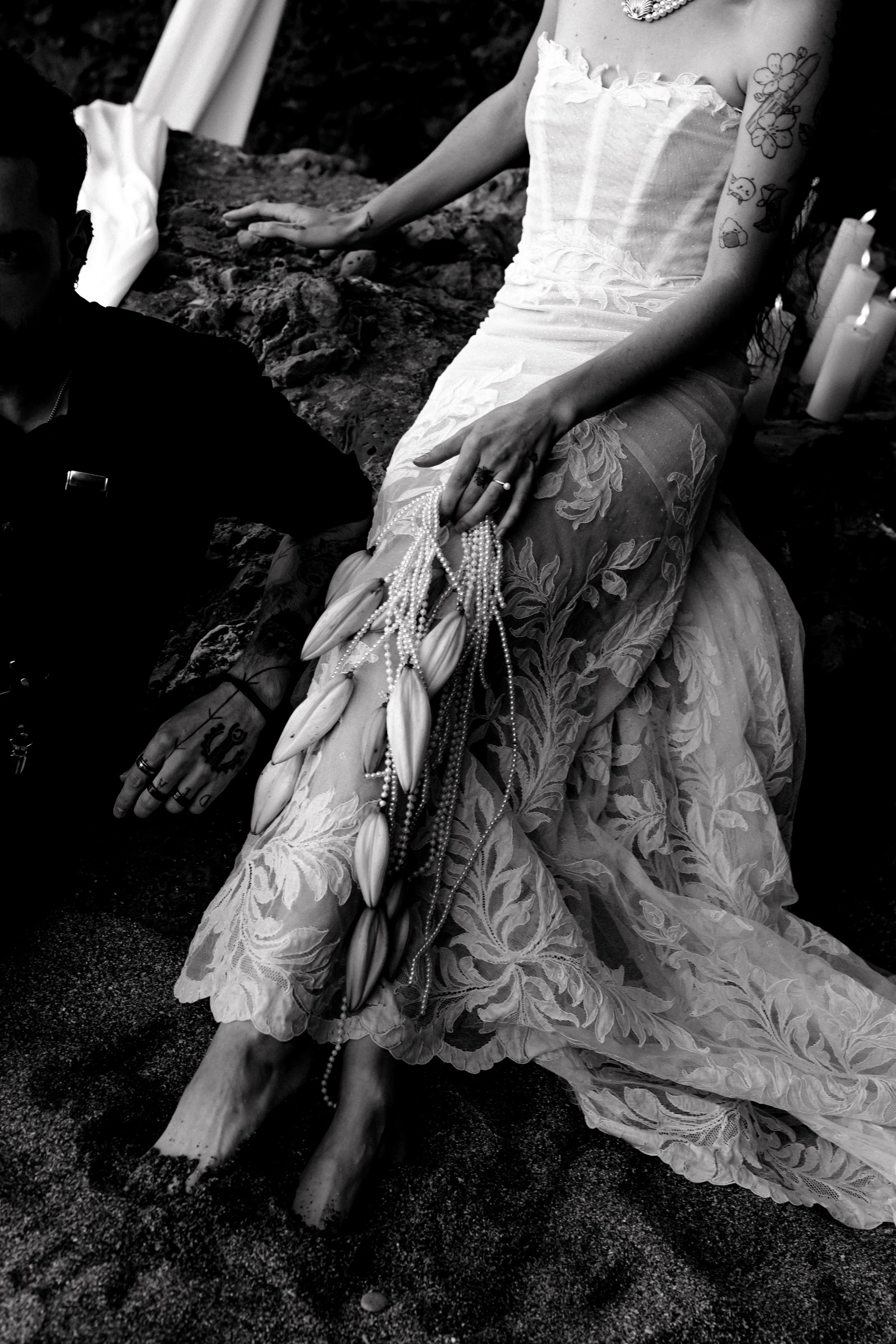 Une femme en robe de mariée, assise avec les pieds dans le sable, tenant un collier de perles et de coquillages, entourée de bougies