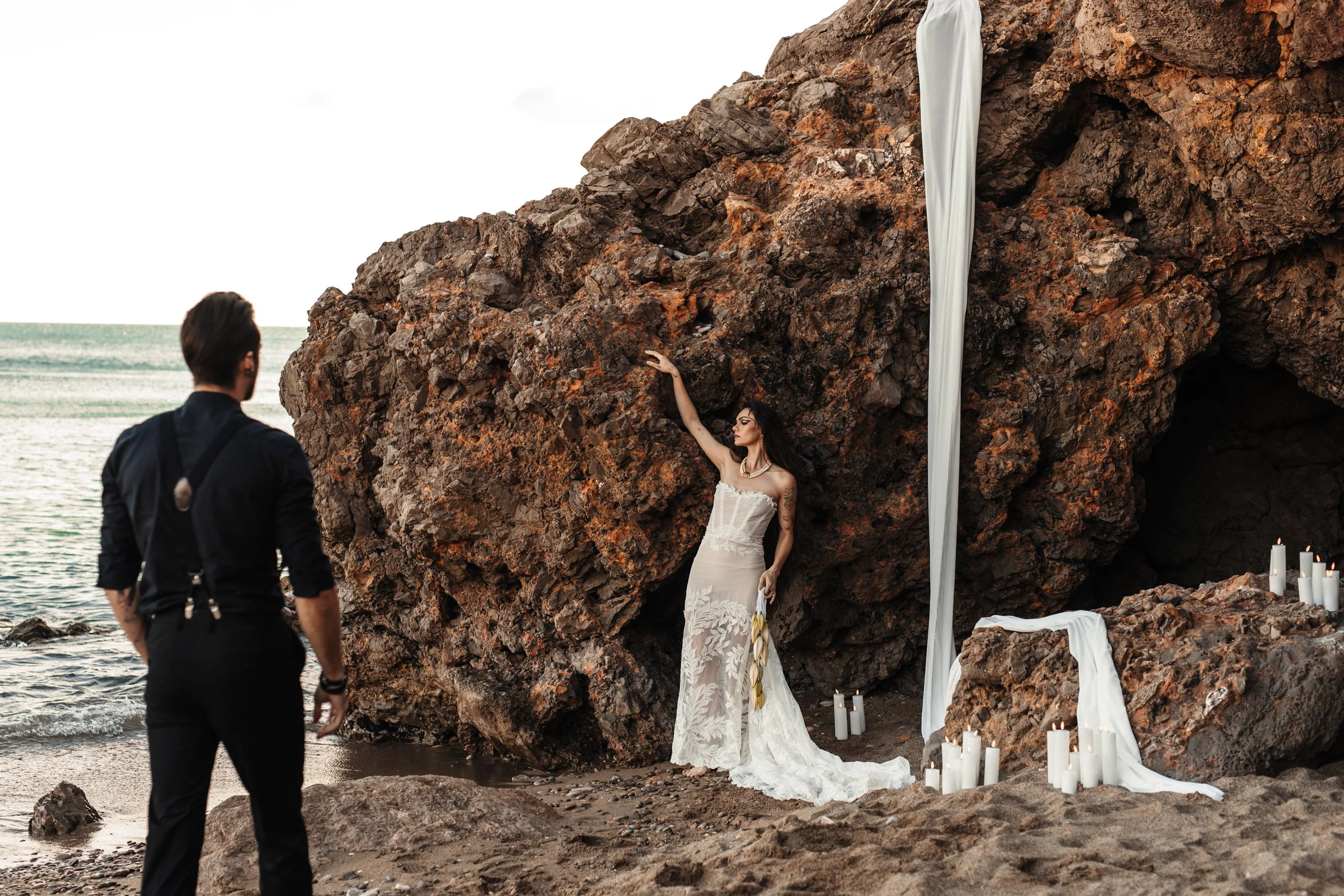 Une femme en robe de mariée se tient devant une roche de la plage avec des bougies blanches et des draps blancs, un homme en tenue noire observe de loin.