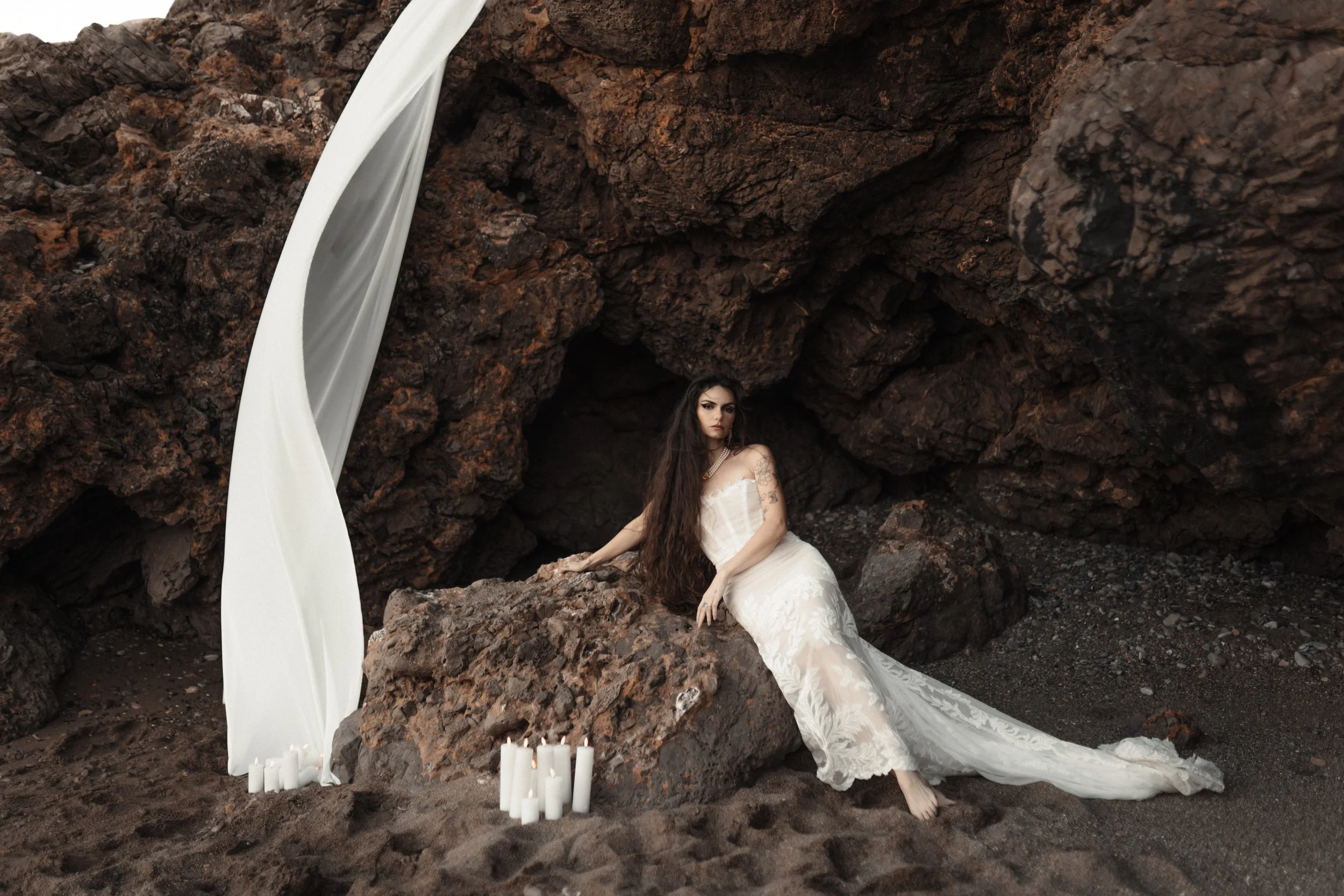 Une femme vêtue d'une robe blanche longue pose sur des rochers noirs près d'une grotte, avec des bougies blanches autour.