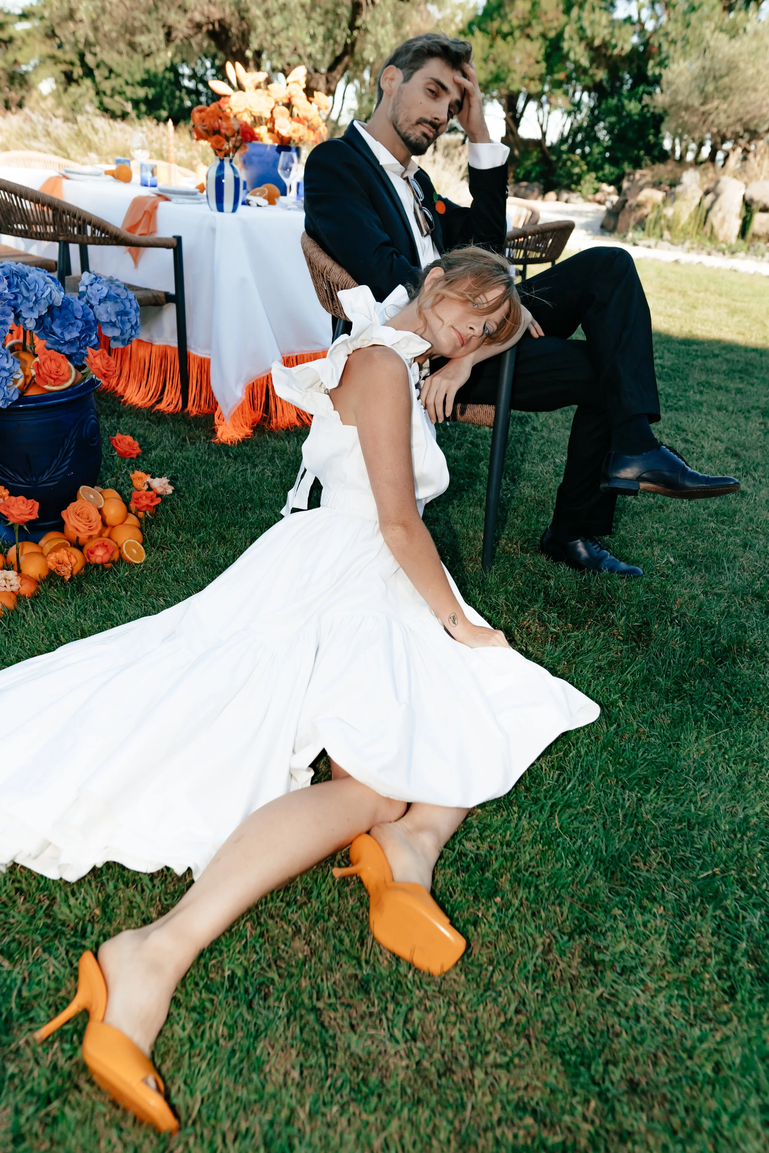 Une jeune femme en robe blanche et chaussures orange allongée sur le sol, apparemment épuisée ou triste, devant une table décorée pour un événement en plein air. Un homme assis derrière elle porte une expression de gêne ou de frustration, avec une ma