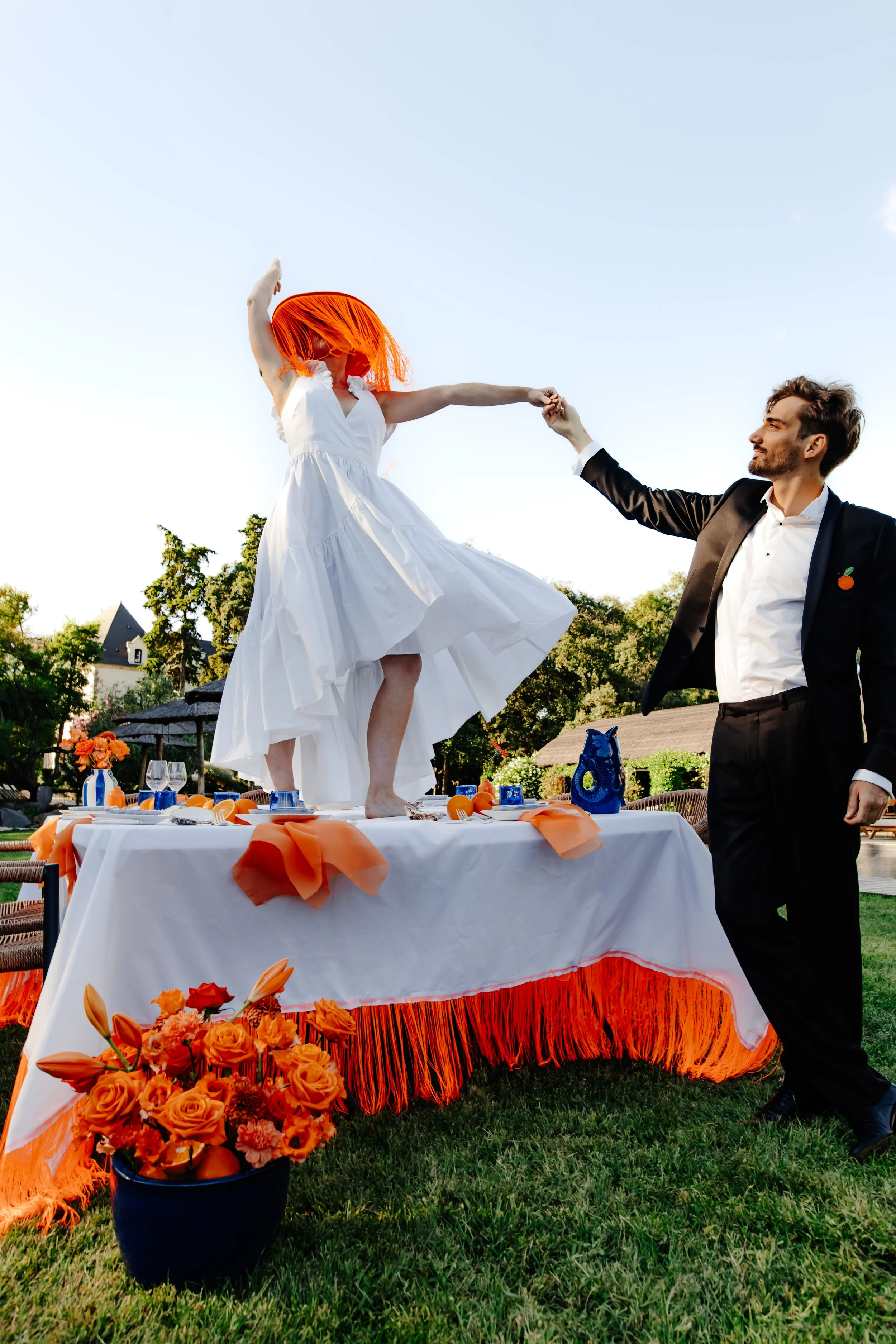 Une femme avec une chevelure orange danse sur une table lors d'une fête en plein air, avec un homme en costume noir qui lui tend la main. La table est décorée de nappes orange et blanche et entourée de fleurs orange.