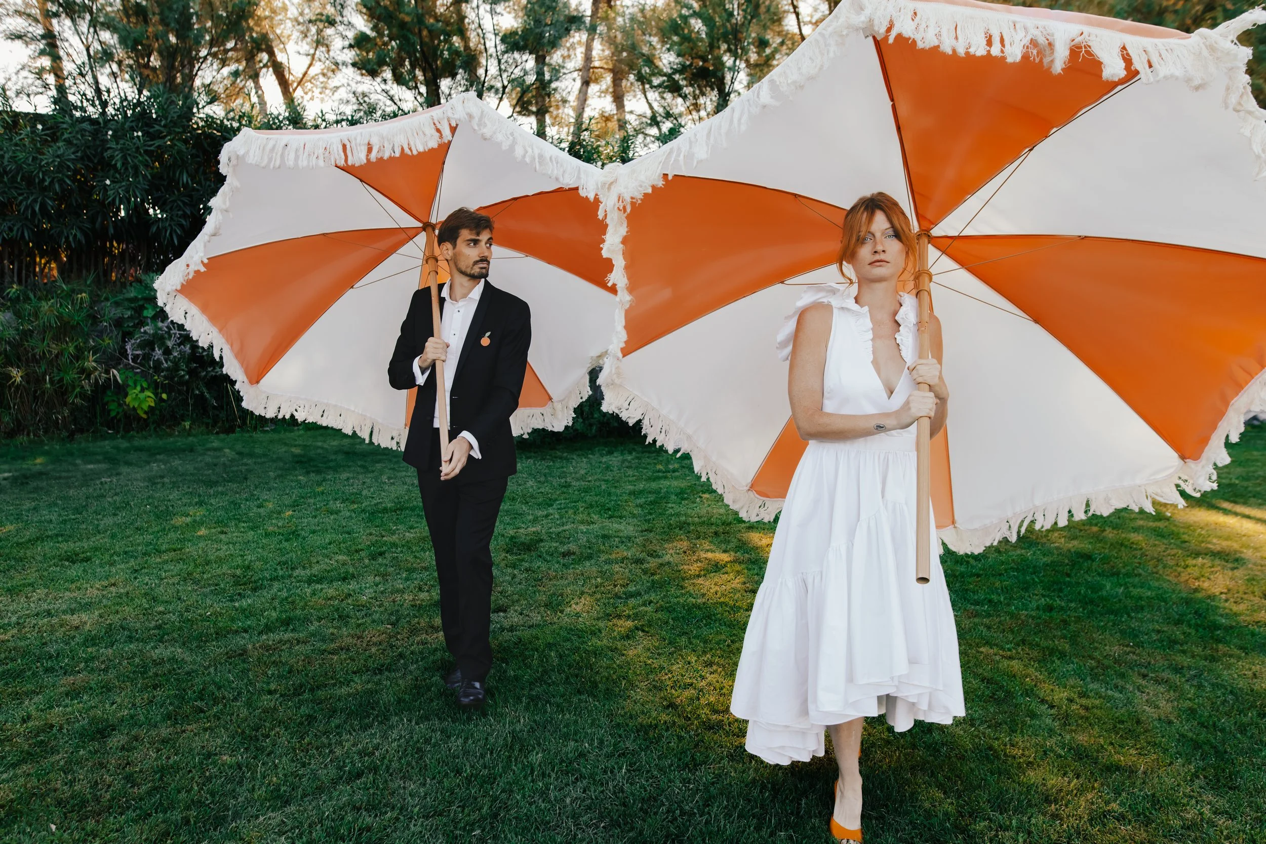 Un homme en costume noir et une femme en robe blanche portent des parapluies orange et blanc dans un jardin.