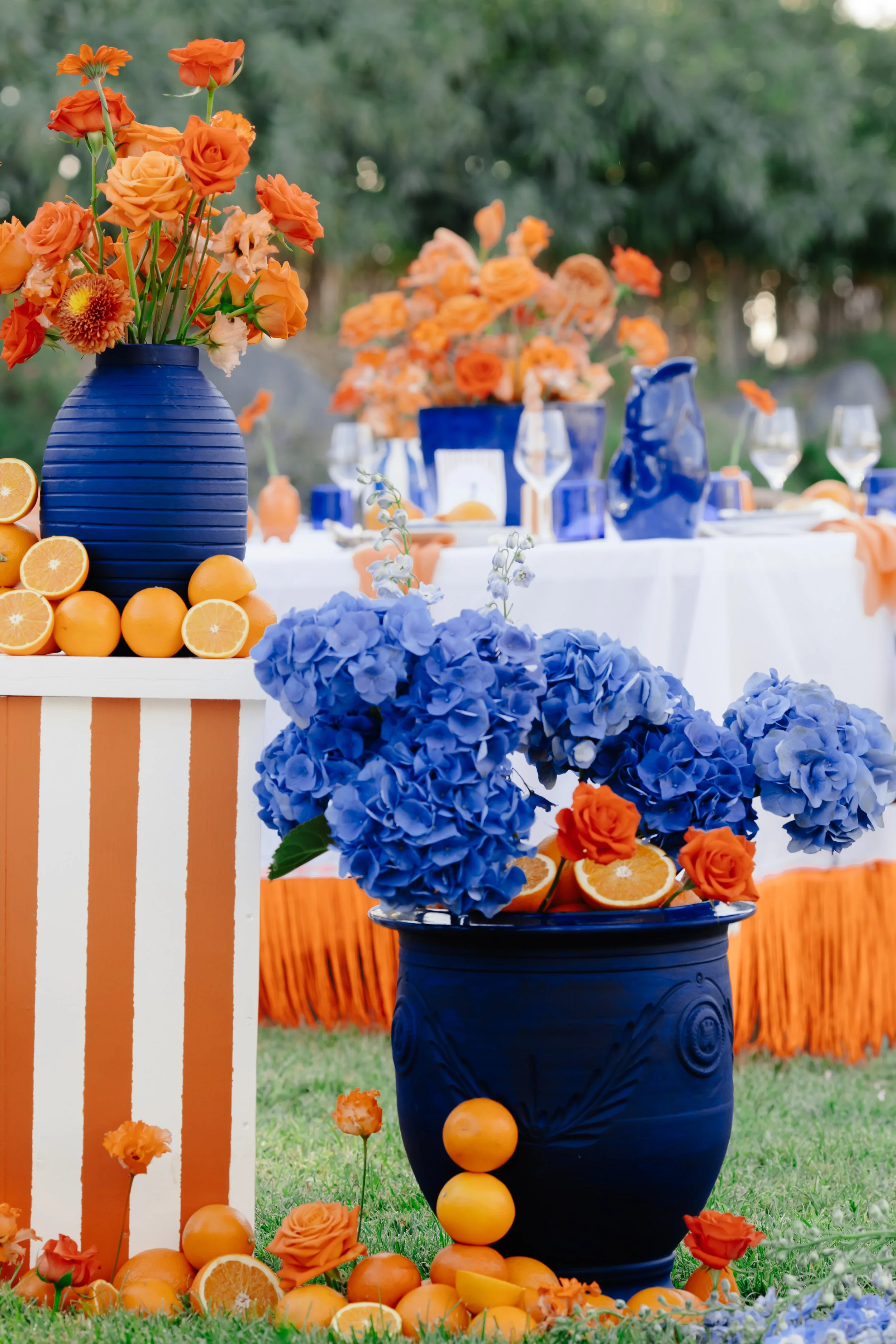 Décor de table extérieur avec des vases bleus contenant des fleurs orange et violet, entouré de oranges, sur un fond d'herbe et de forêt.