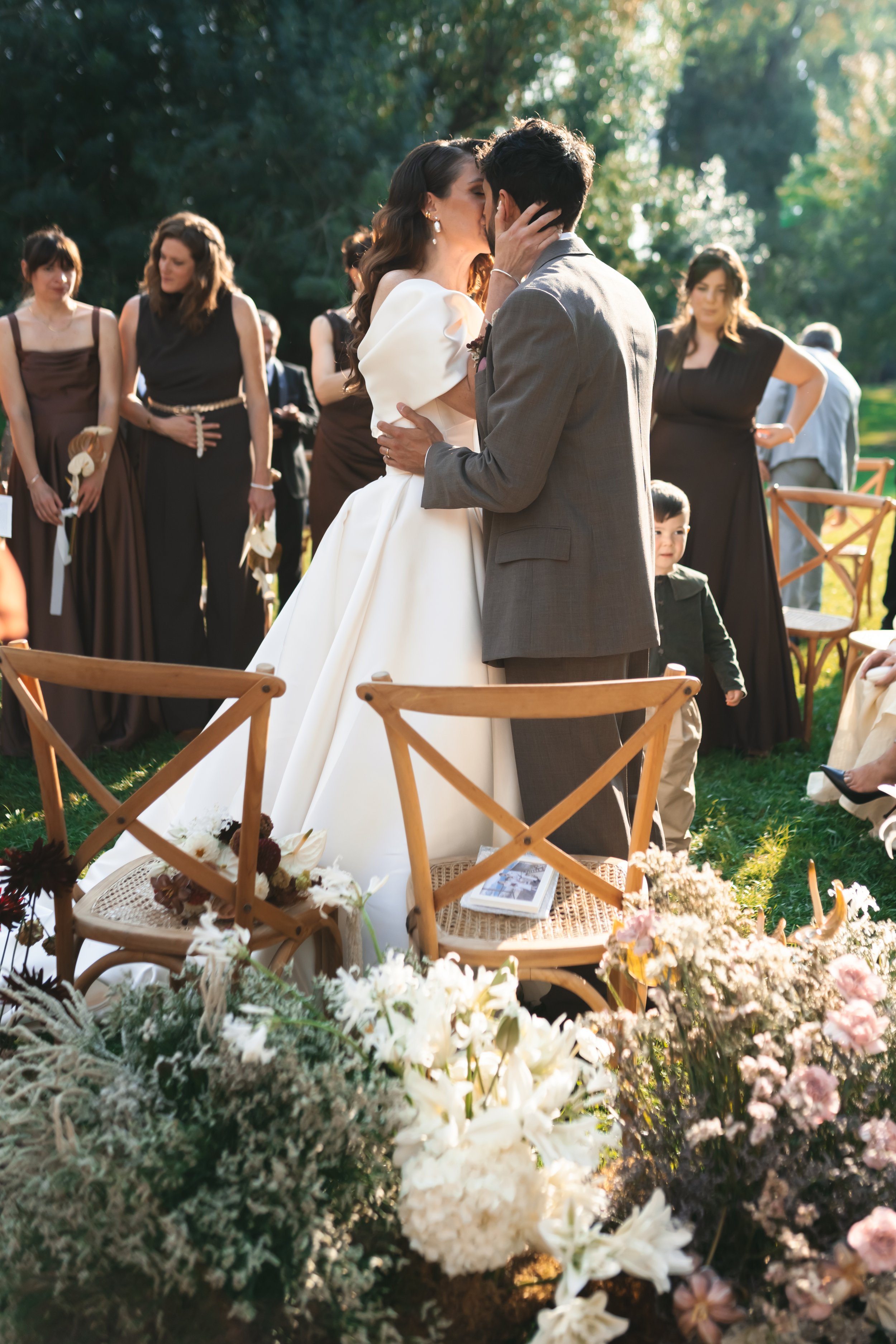 Un couple en costume de mariage s'embrasse lors d'une cérémonie en plein air, entourés d'invités et de fleurs.