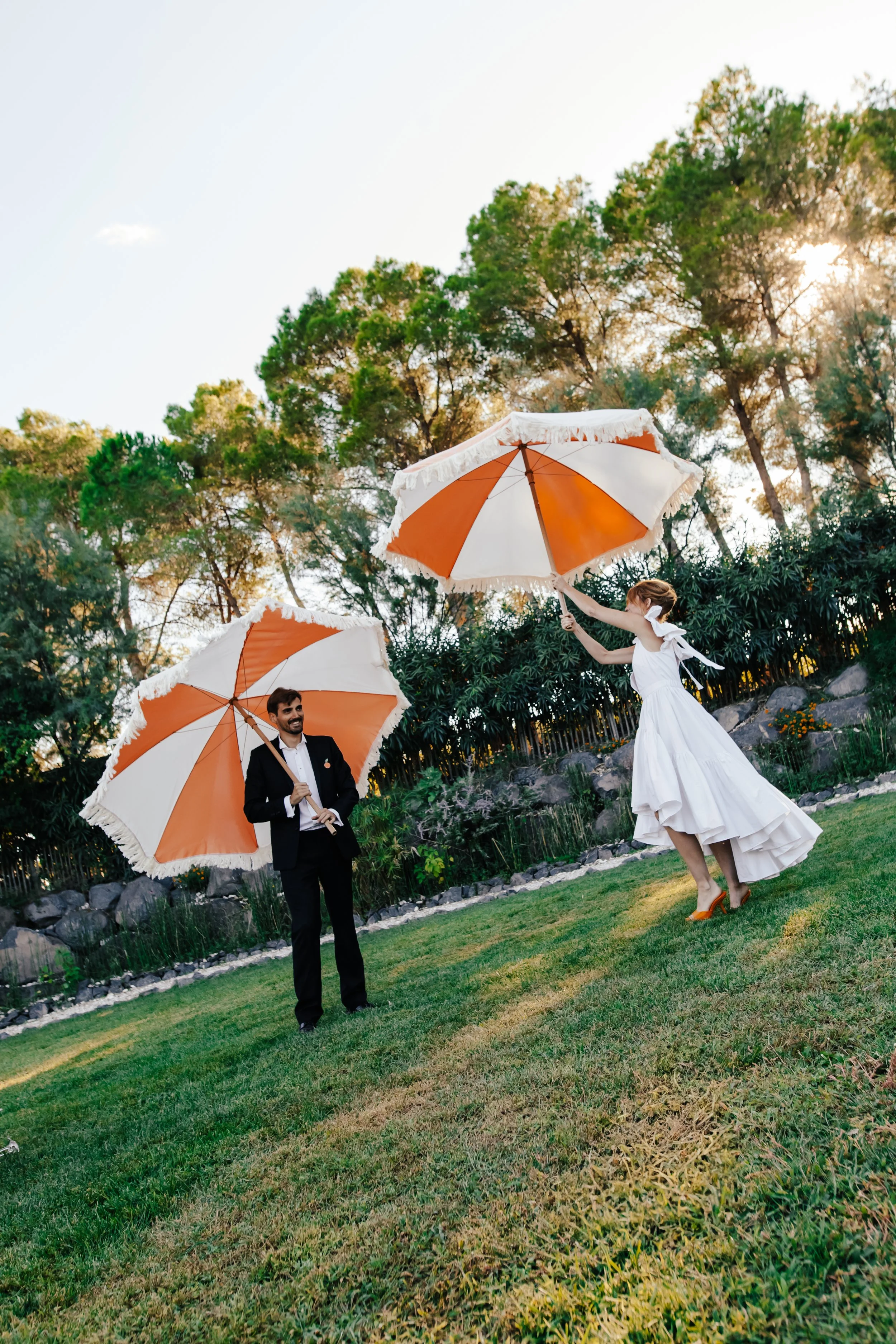Un couple habillé élégamment dans un jardin, chacun tenant un parasol orange et blanc, sous un ciel ensoleillé.