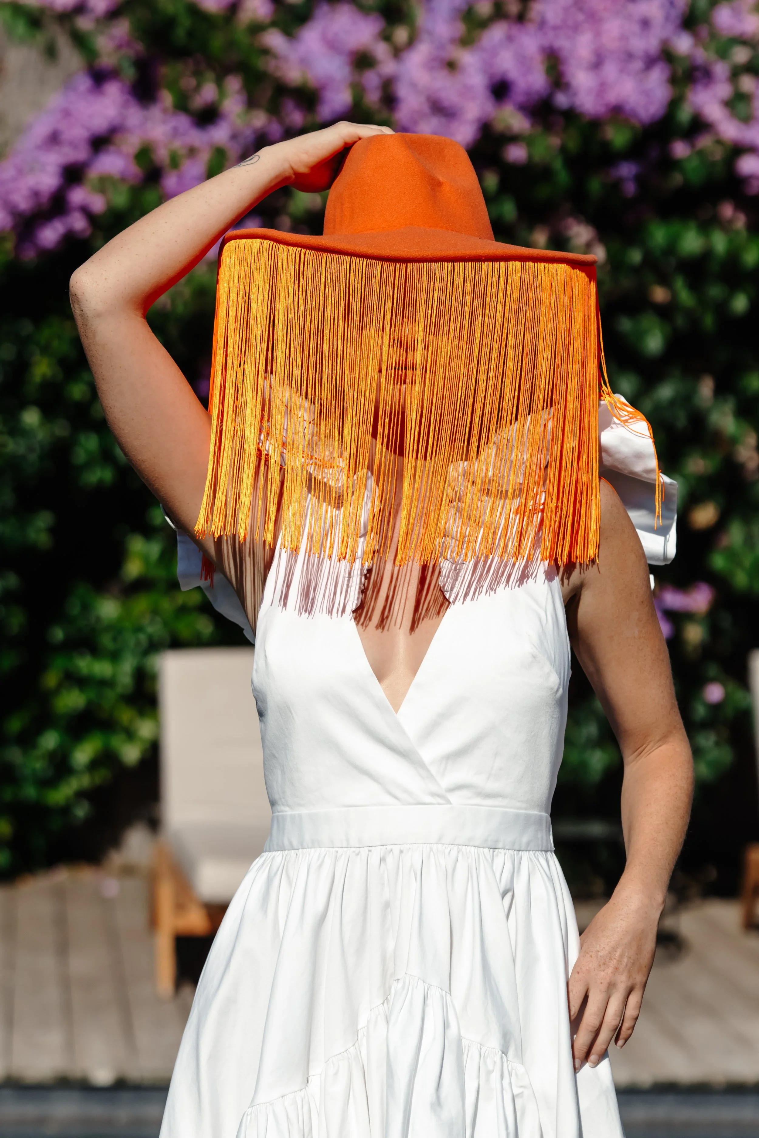 Une femme portant un chapeau orange avec un voile à franges oranges, une robe blanche avec des épaulettes en tissu blanc, debout devant un arrière-plan de fleurs violettes et de végétation