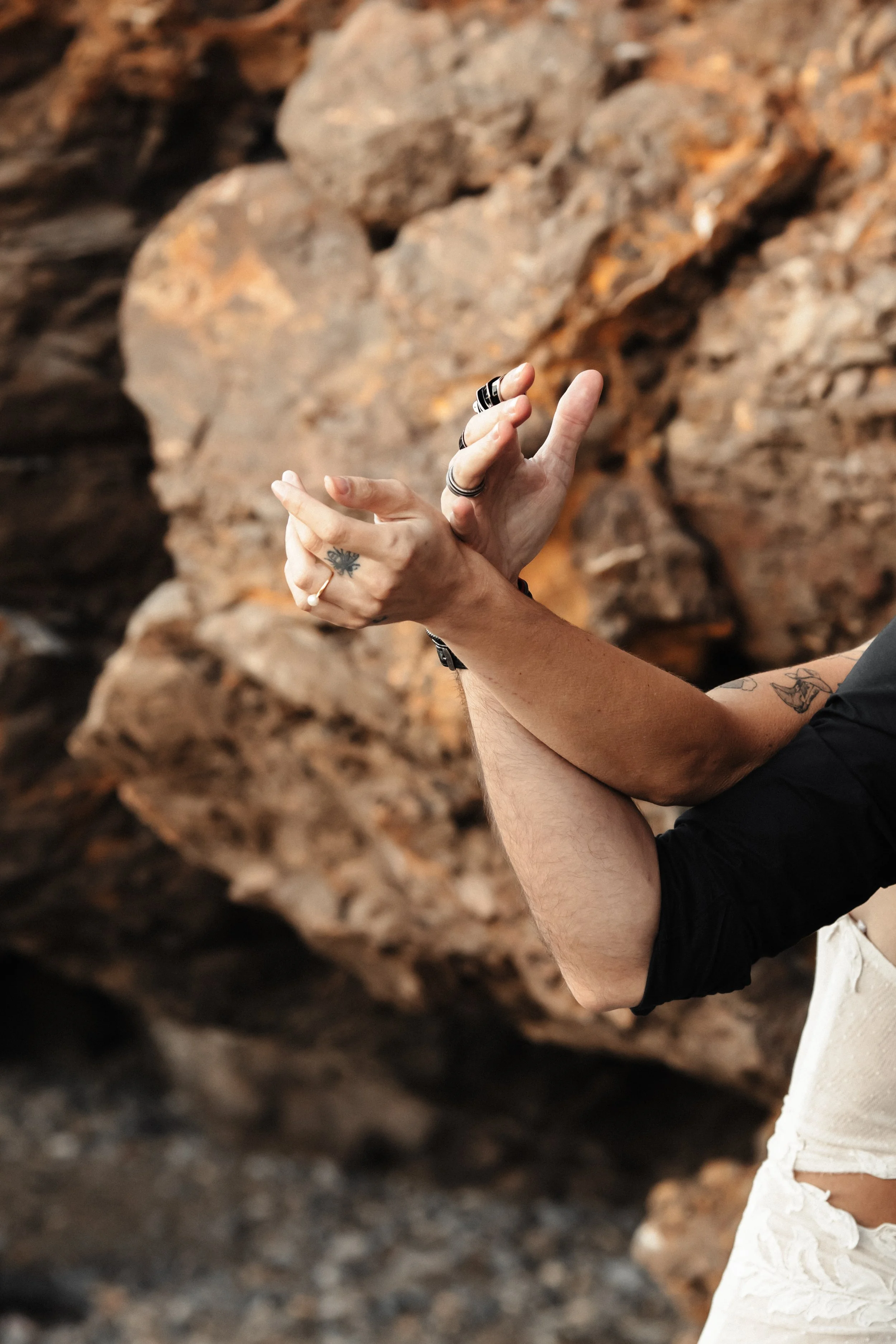 Mains croisées avec bijoux et tatouages, contre un fond rocheux.