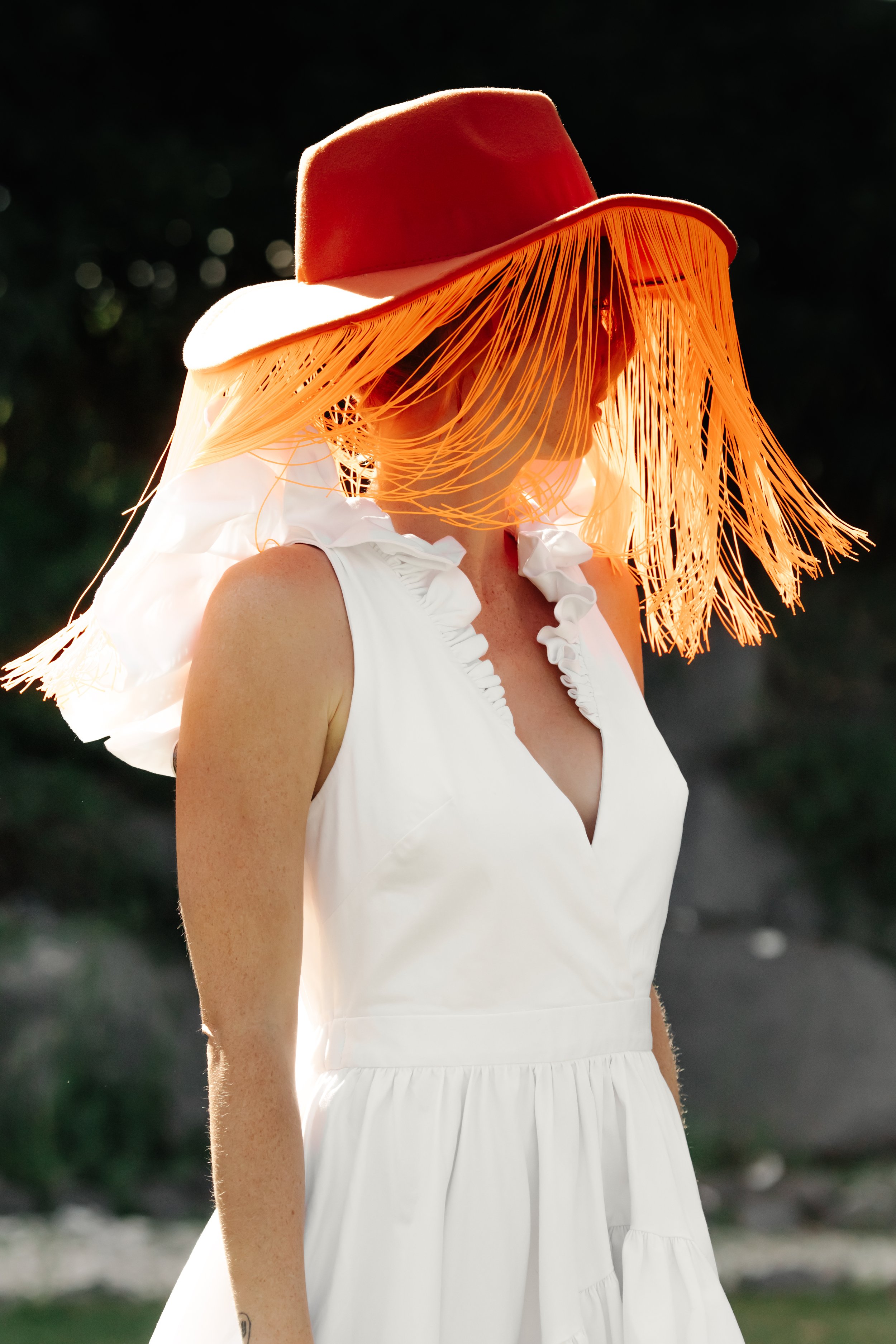 Femme portant un chapeau rouge avec des franges en plastique, habillée d'une robe blanche avec des bretelles froncées, en plein air dans un environnement naturel.