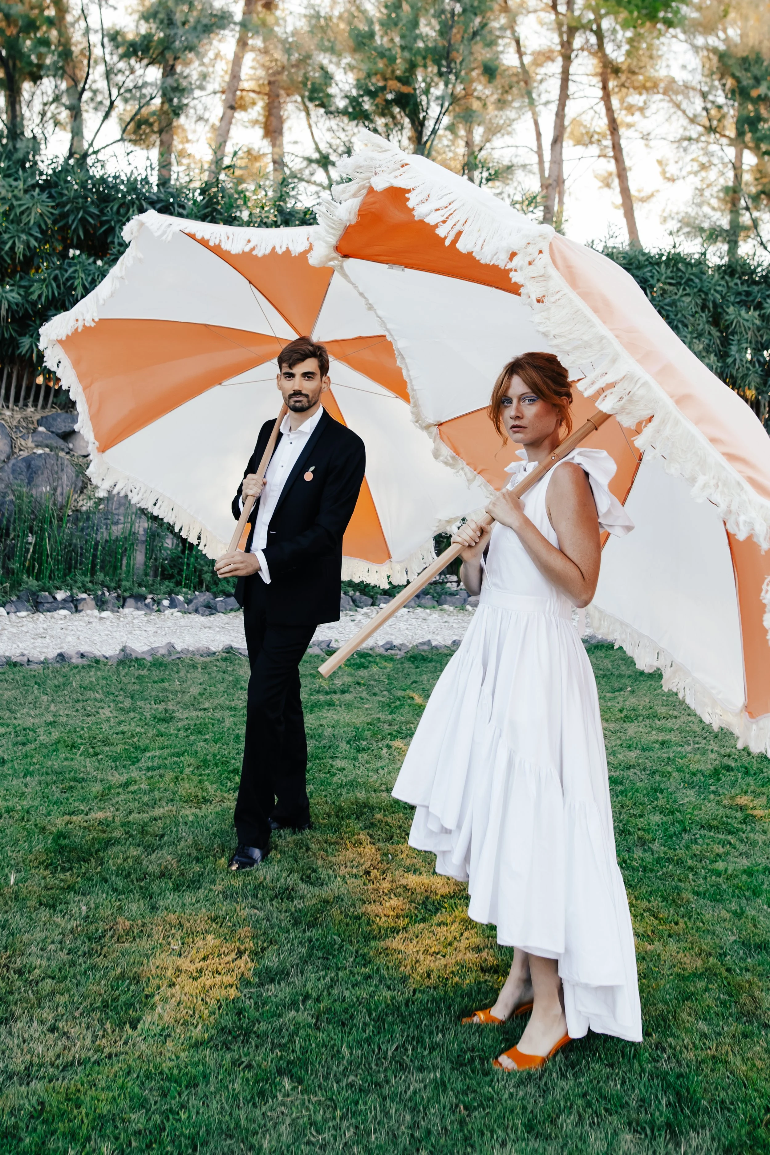 Un homme et une femme se tiennent dans un jardin, chacun tenant une grande parasol beige et orange avec des franges blanches, tous deux vêtus élégamment, la femme en robe blanche et le homme en costume noir.