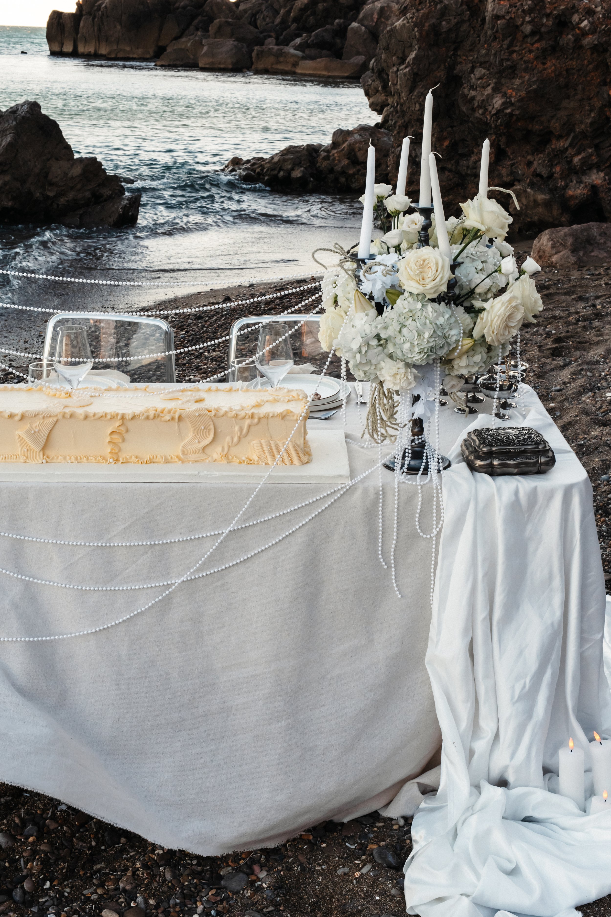 Table de mariage décorée avec un bouquet de fleurs blanches, des bougies, des candélabres, et des perles en plastique, située sur une plage rocheuse avec vue sur la mer.