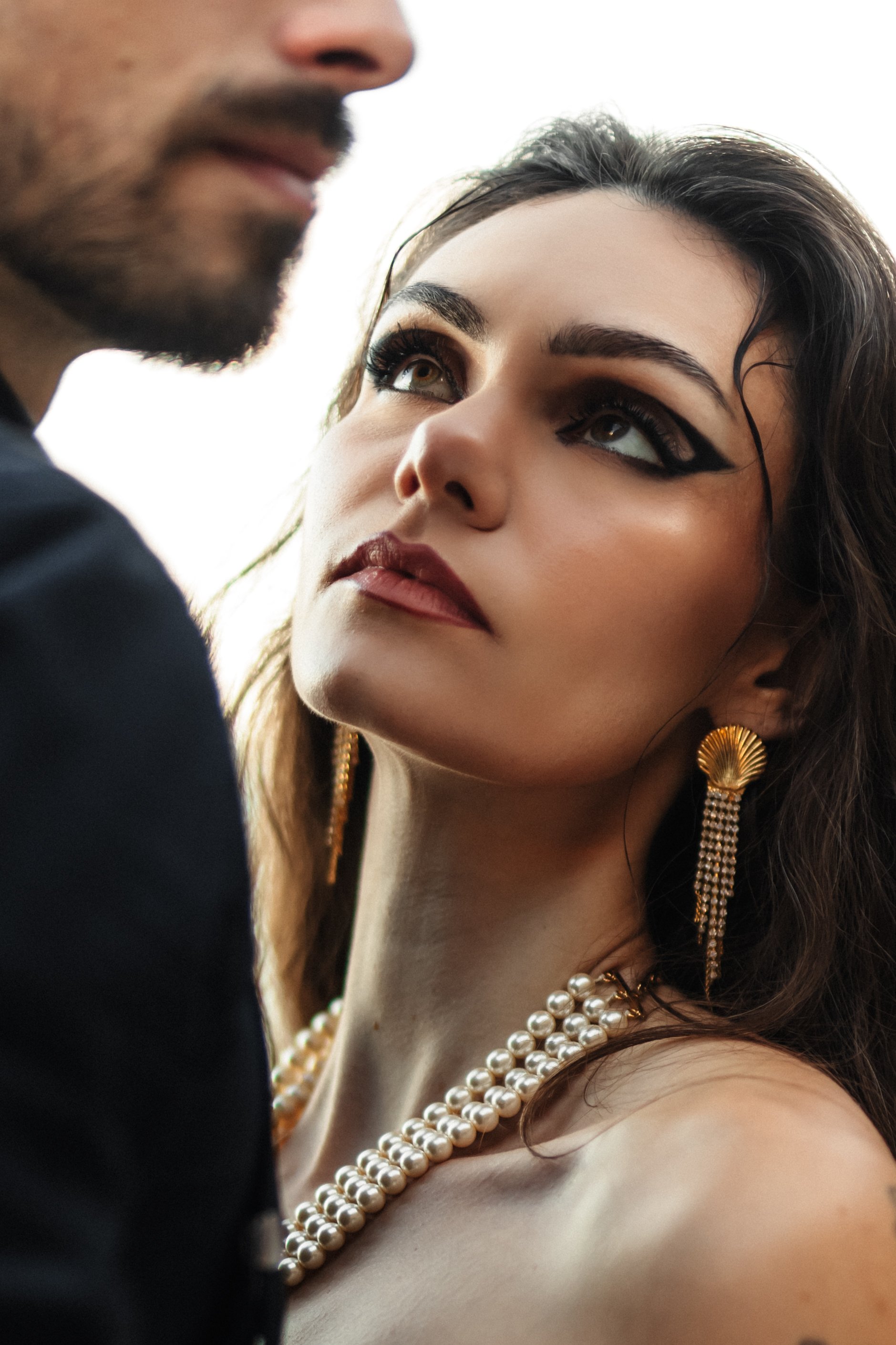 Une femme avec des bijoux en perles, yeux maquillés et bijoux dorés, regarde un homme dont le visage est partiellement visible.
