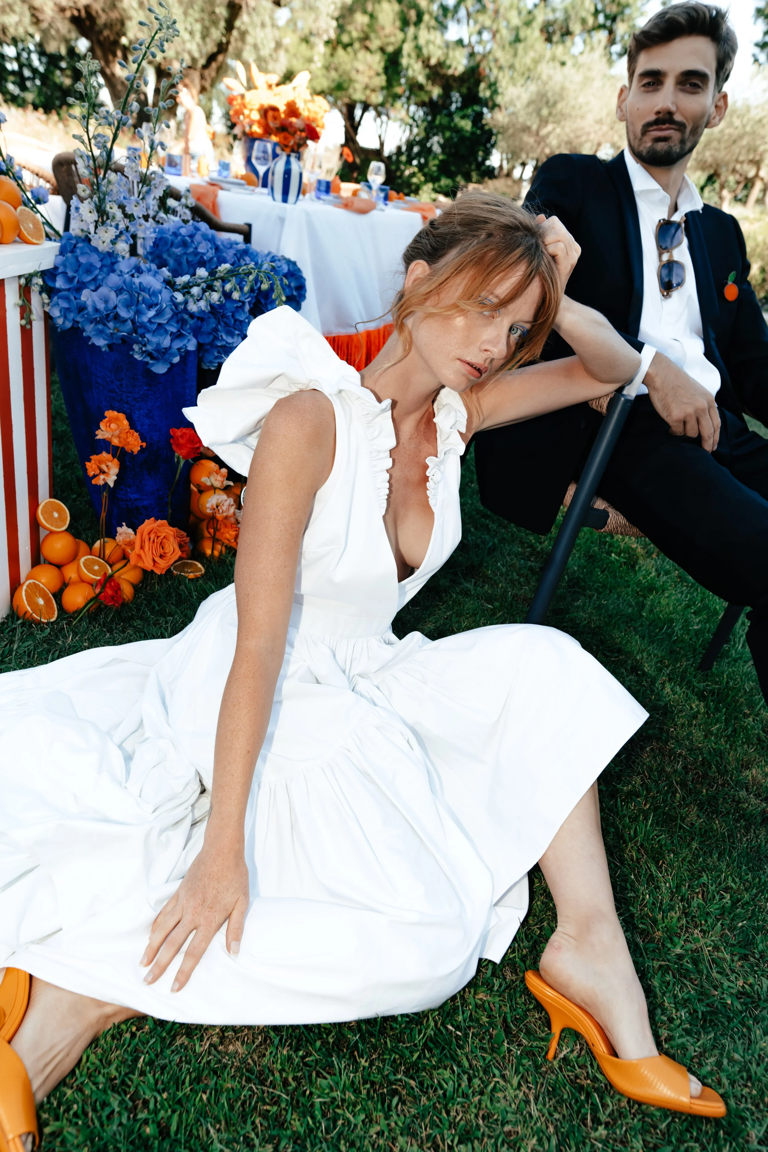Une femme en robe blanche et des talons oranges, assise sur l'herbe à côté d'une table décorée de fleurs et d'agrumes, avec un homme en costume derrière elle lors d'une fête en plein air.