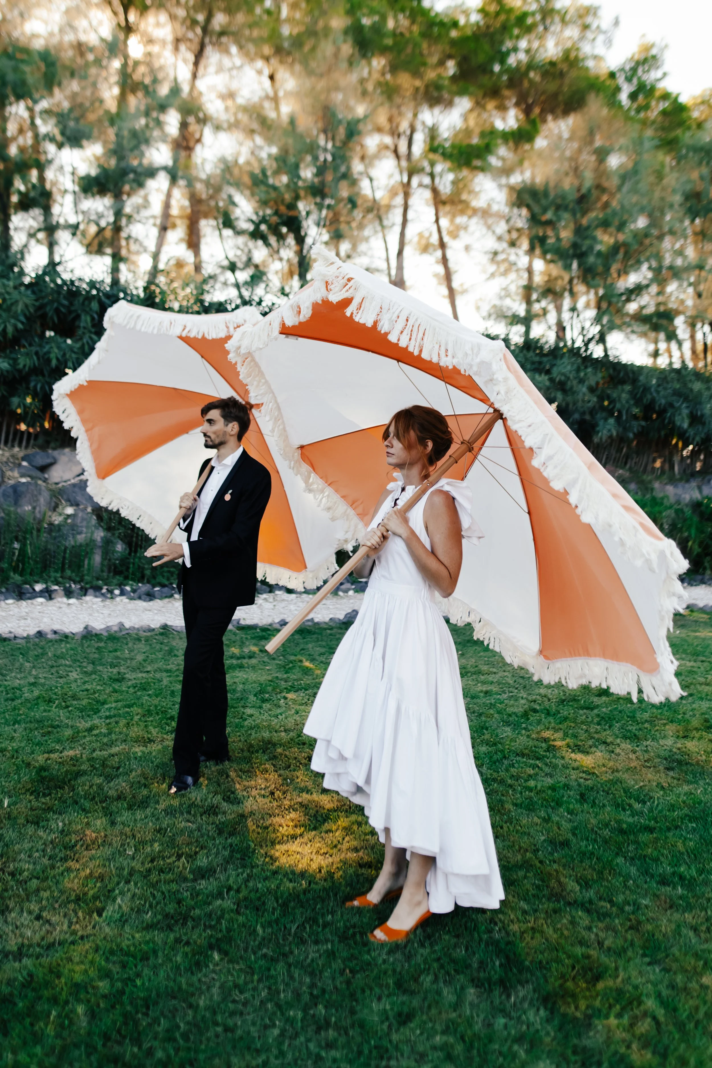 Un homme en costume noir et une femme en robe blanche, tenant des parapluies orange et blanc avec des bordures en franges, marchent sur de l'herbe dans un parc.