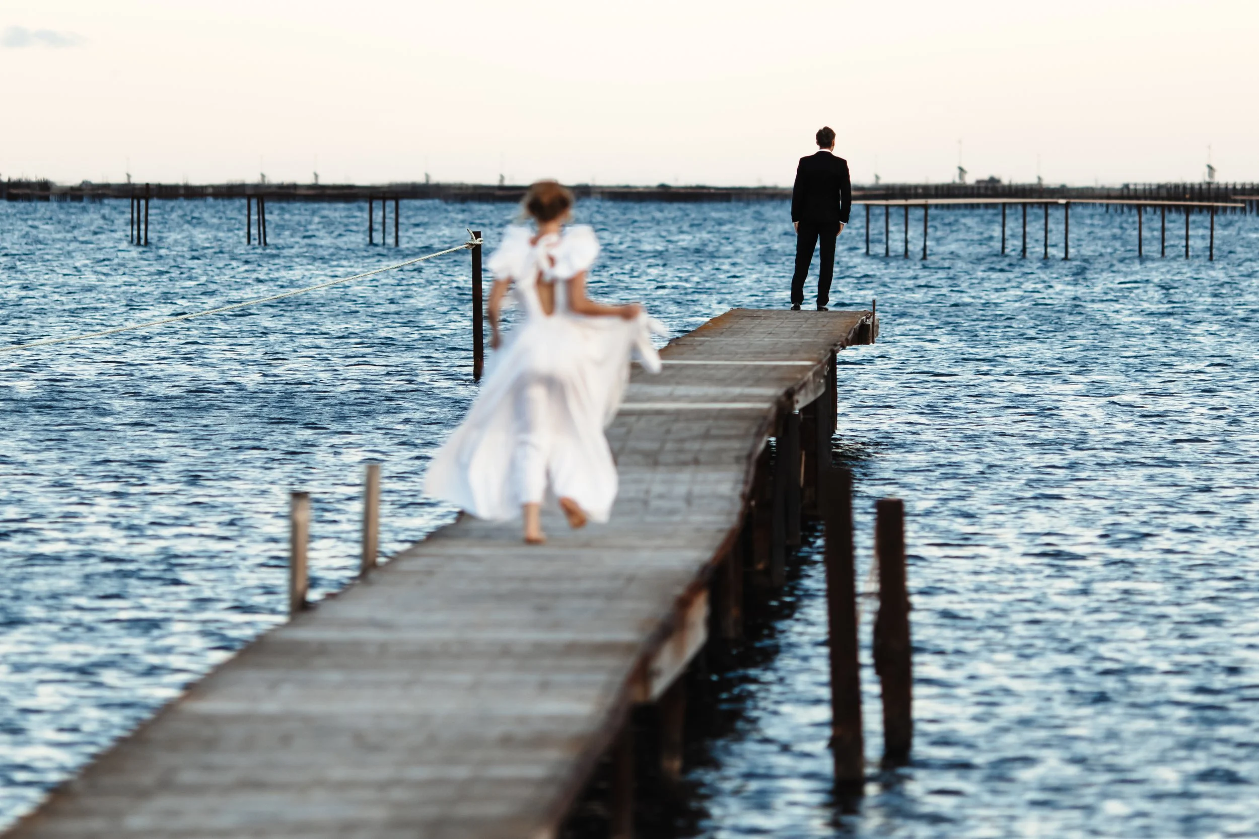 Une femme en robe blanche et un homme en costume noir sur une jetée en bois au bord de l'eau, avec une horizon au coucher de soleil.
