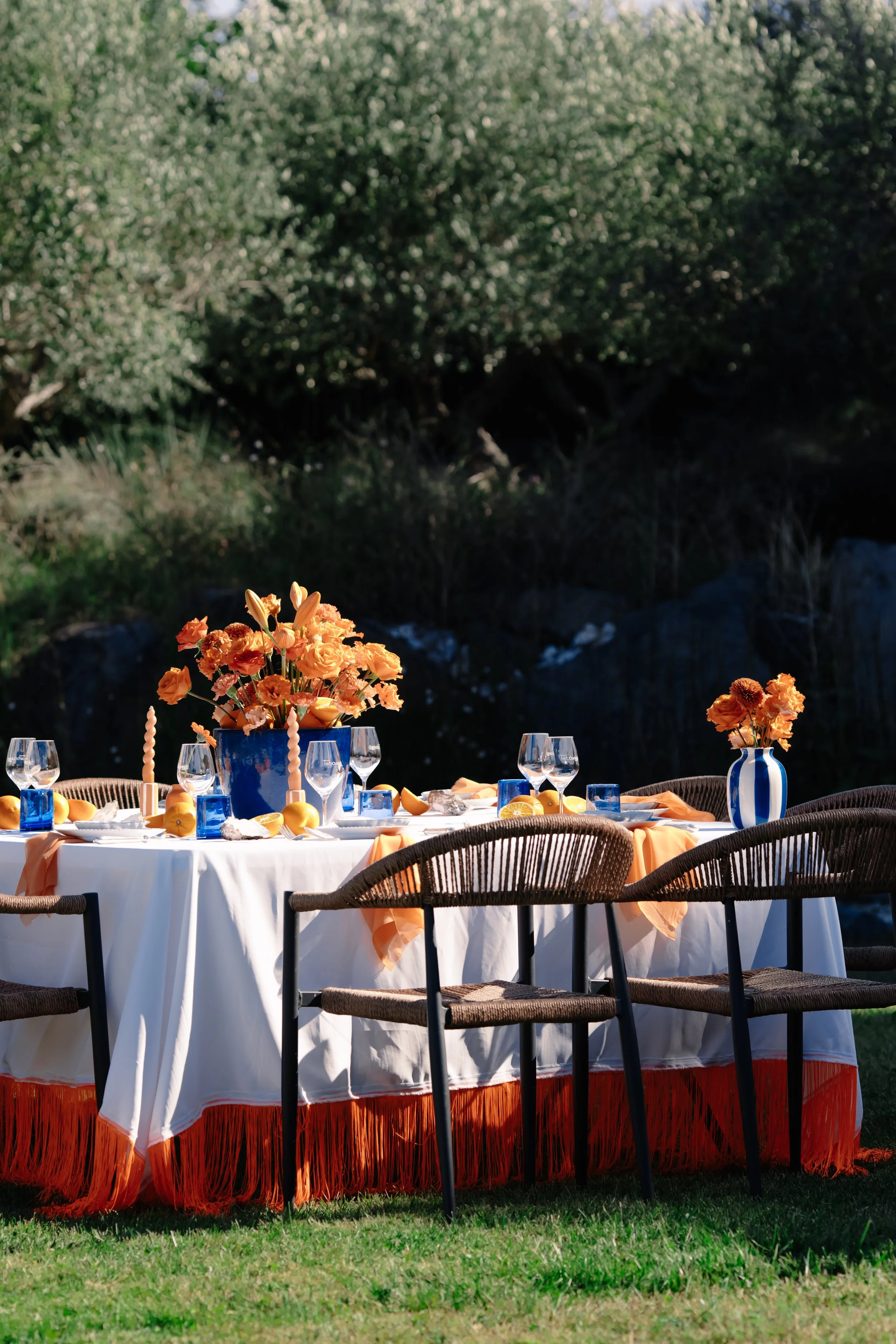 Table de repas en plein air, décorée avec des fleurs orange dans des vases bleus et blancs, des chandeliers, des assiettes, des verres à vin et des citrons, sous un ciel ensoleillé.