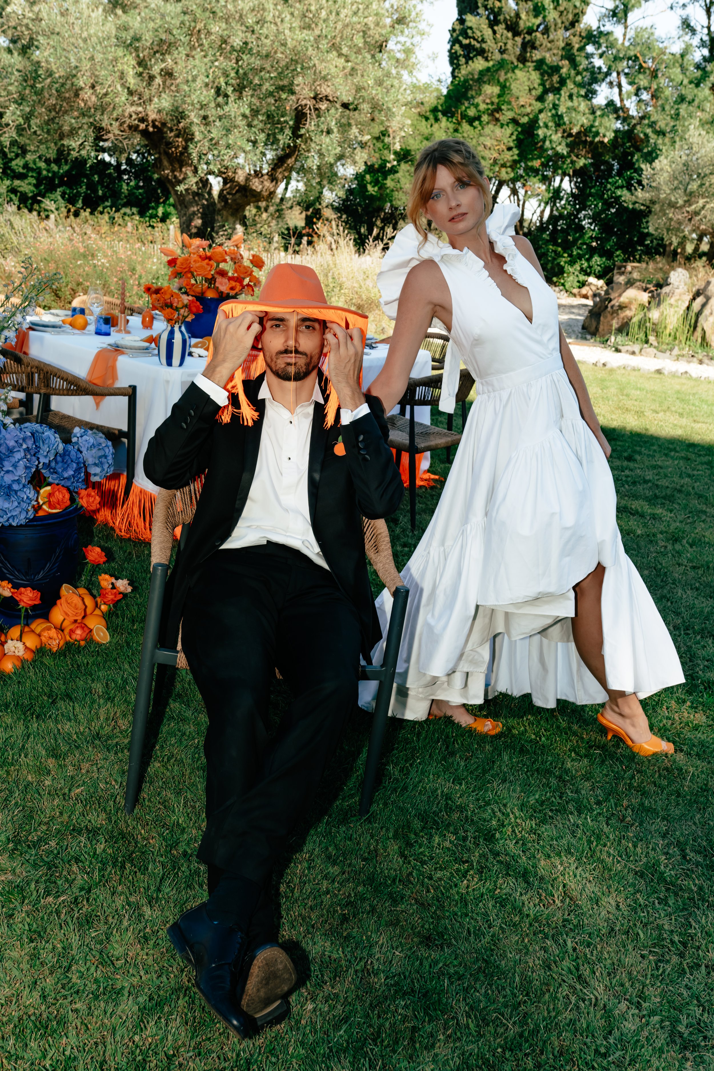 Un homme et une femme posent lors d'une fête en plein air. L'homme porte un costume noir et un chapeau orange, tandis que la femme porte une robe blanche longue et des chaussures orange. La table de fête décorée en orange et bleu est visible en arriè