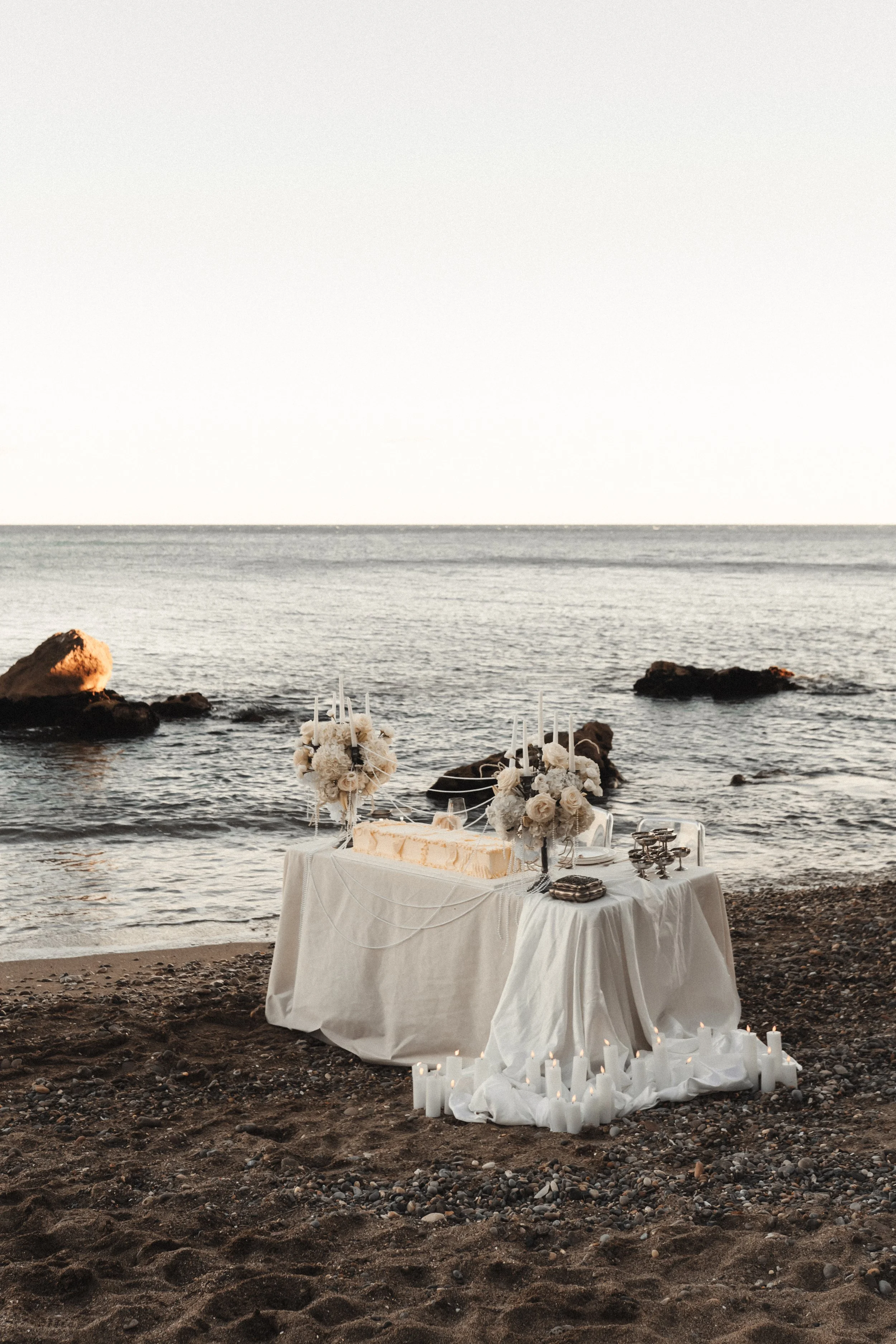 Table dressée pour un mariage ou une cérémonie sur une plage, décorée avec des fleurs, des chandelles et un gâteau, avec l'océan en arrière-plan.