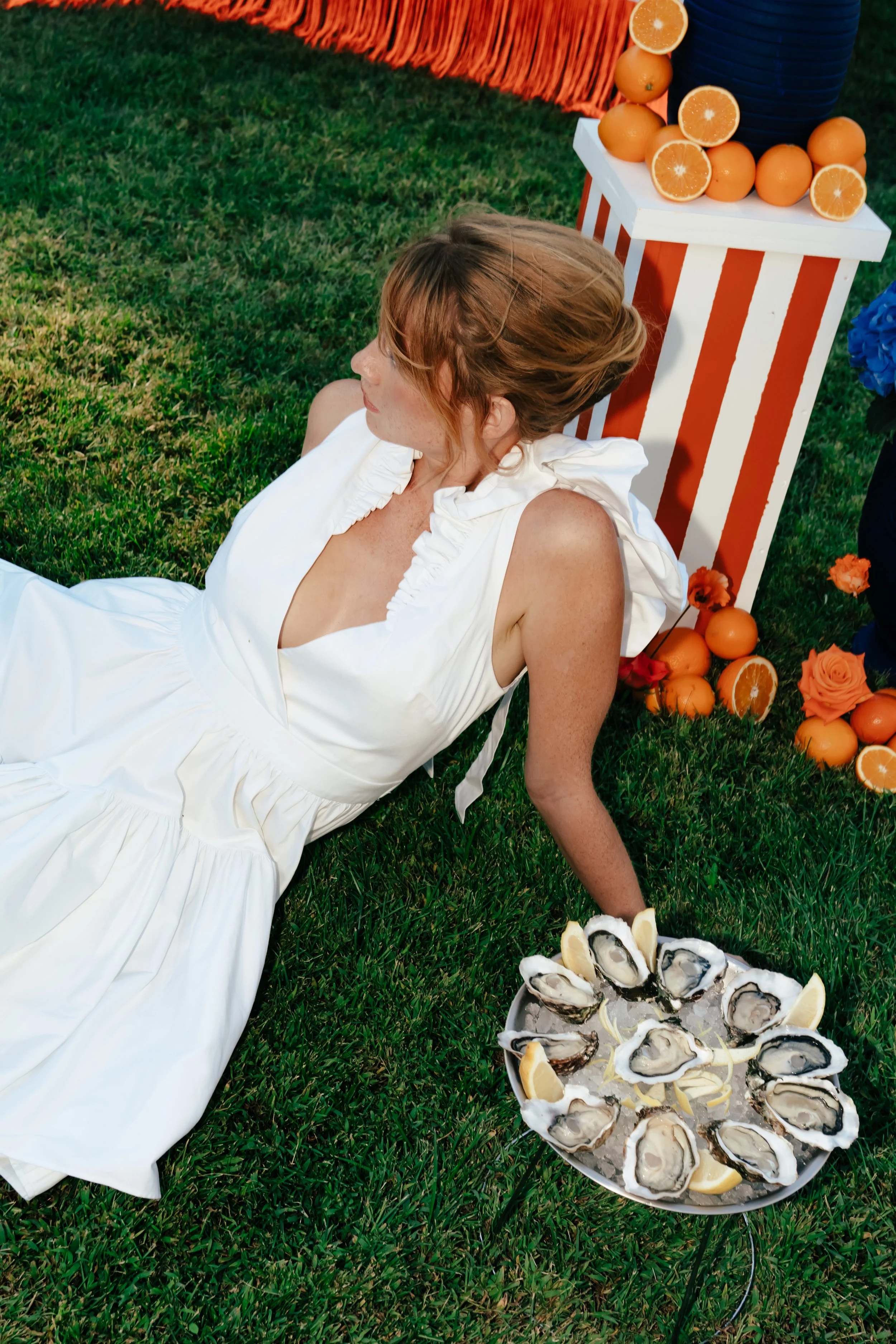 Femme en robe blanche, allongée sur l'herbe, près d'une assiette d'huîtres garnie de quartiers de citron, avec un stand décoré d'oranges et de fleurs colorées en arrière-plan.