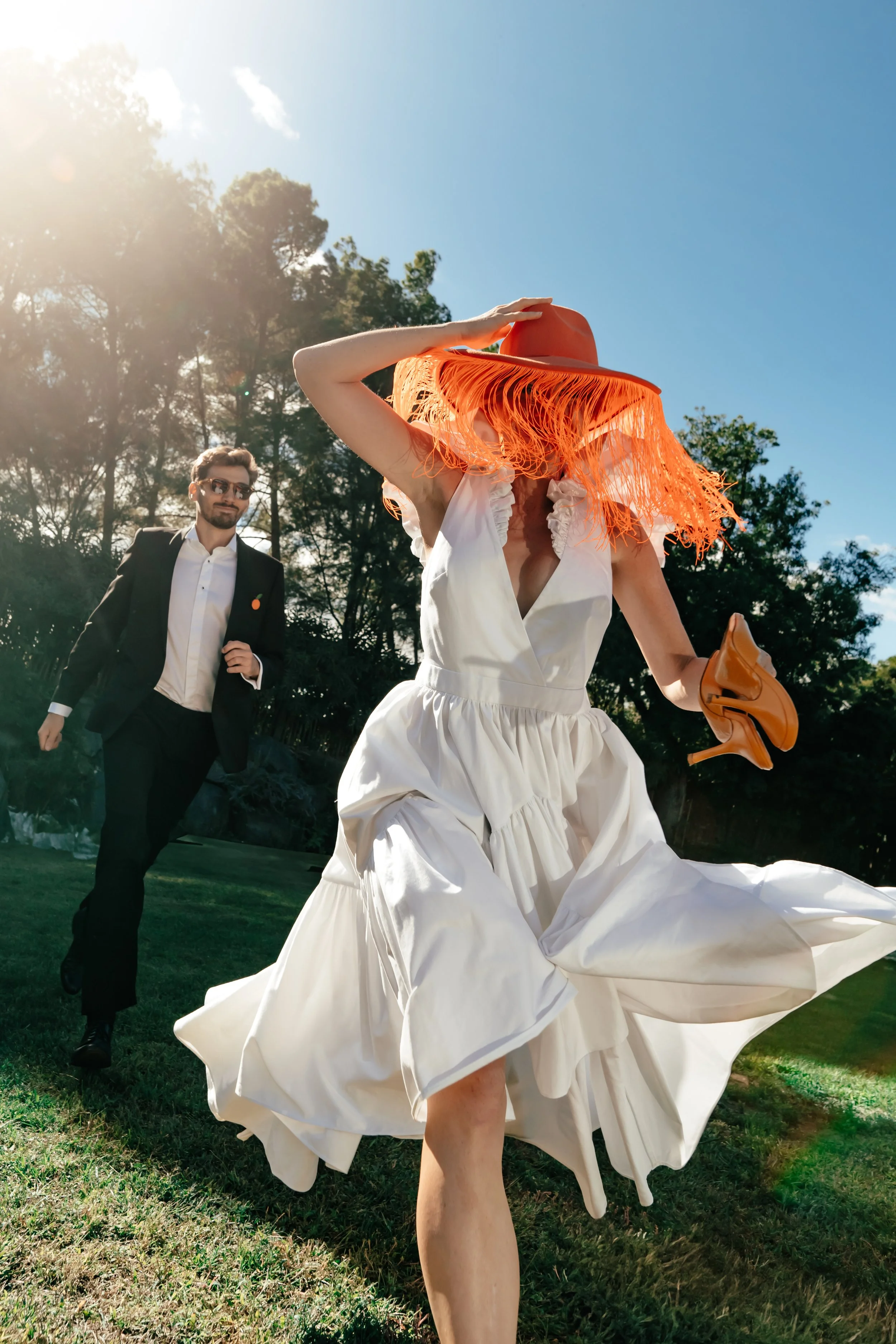 Une femme en robe blanche et chapeau orange avec des franges qui court dans un jardin, avec un homme en costume noir et lunettes derrière elle, en plein soleil.