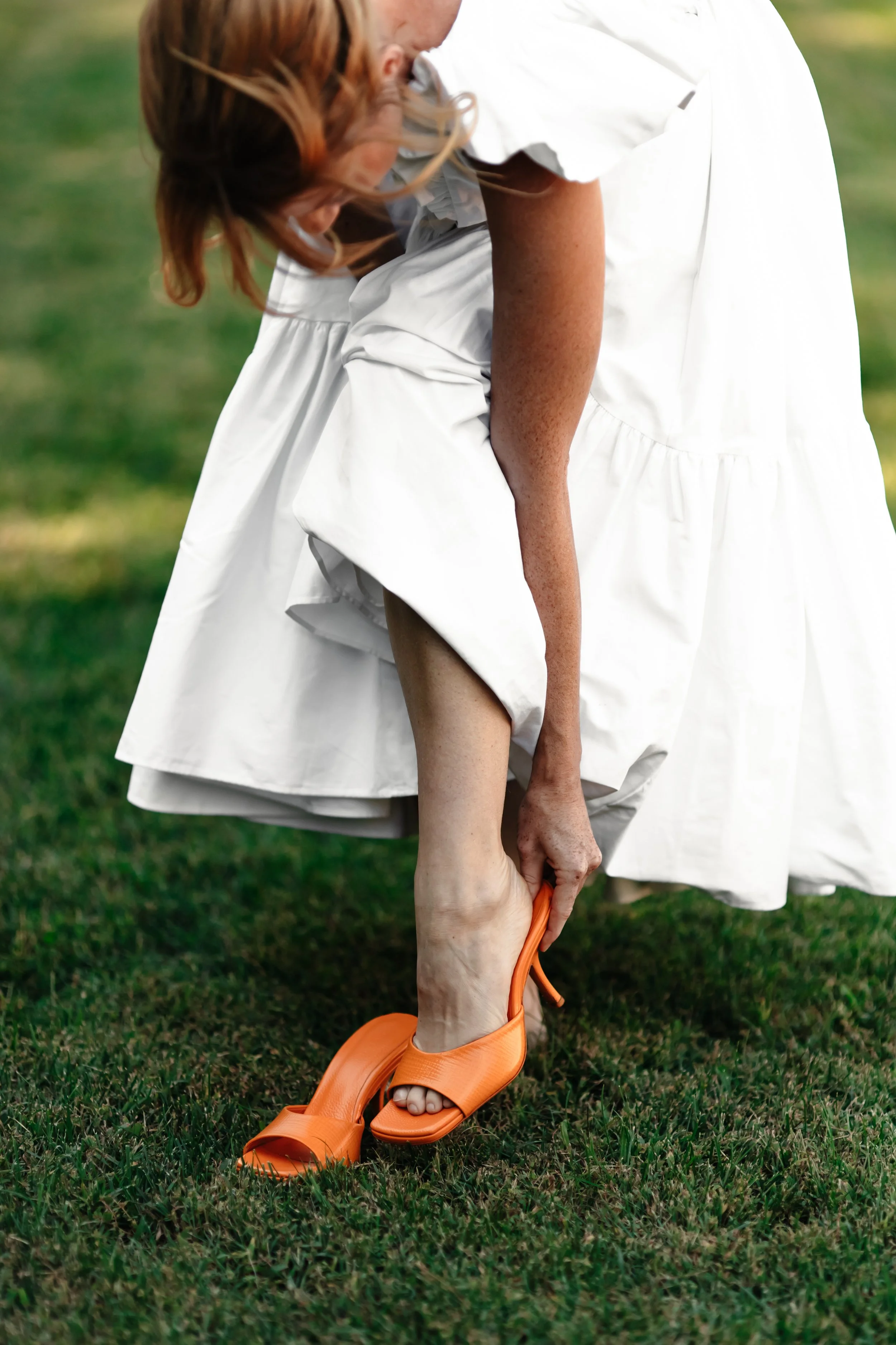 Une femme en robe blanche ajuste sa chaussure à talons orange sur l'herbe.