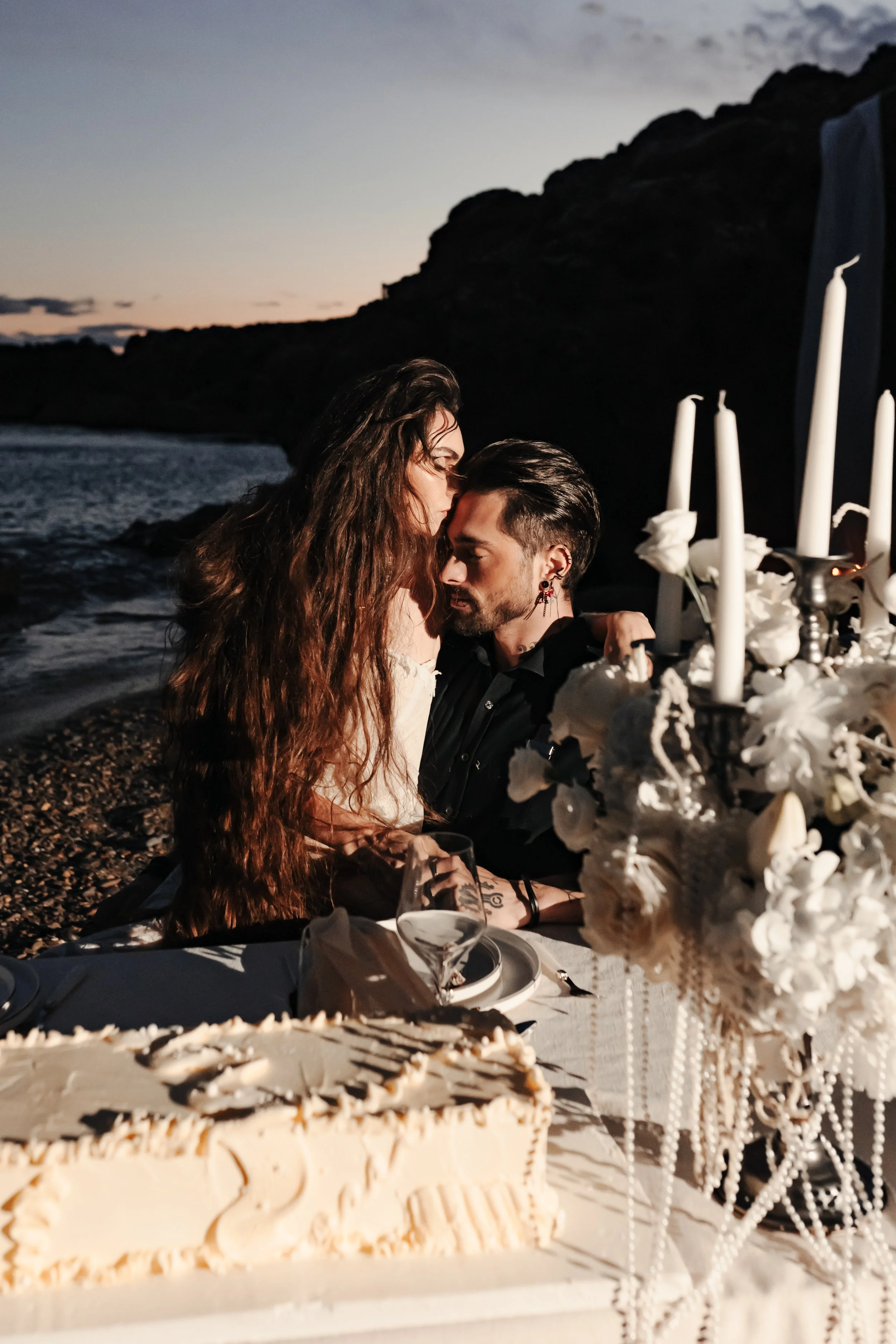 Un couple lors d'un dîner romantique sur la plage au coucher du soleil, entouré de décorations blanches, avec un gâteau et des chandeliers.