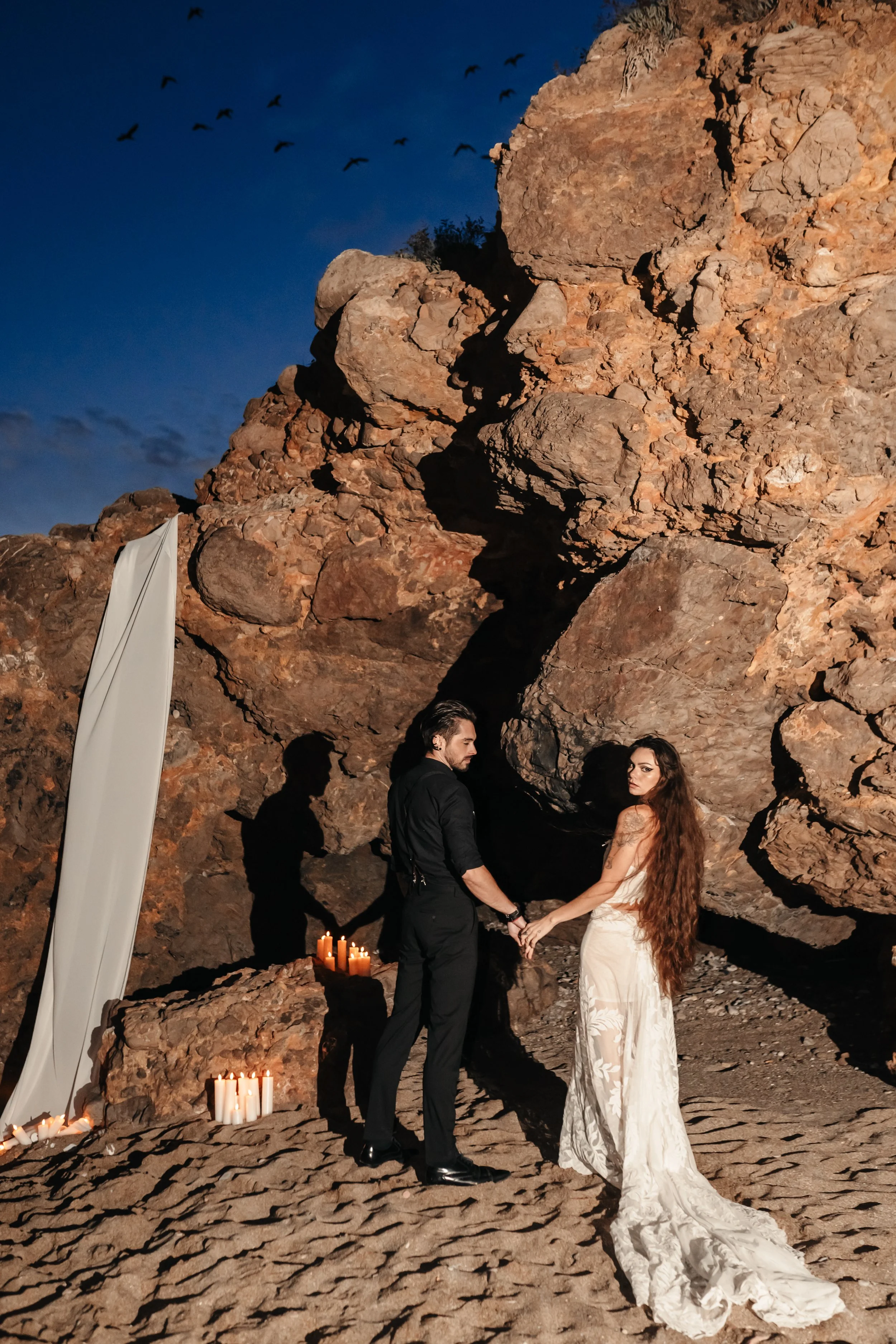Un couple en tenue de mariage se tenant la main devant des rochers, avec des bougies allumées à proximité et un ciel sombre en soirée.