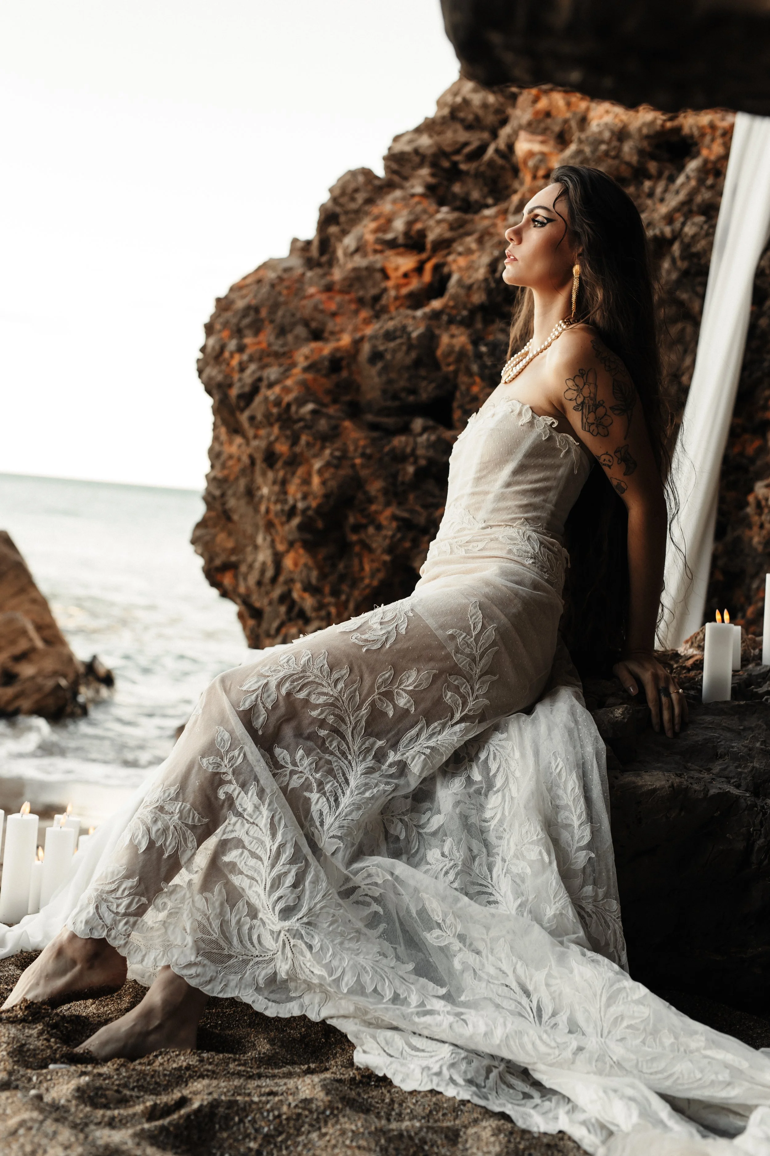 Une femme en robe de mariée blanche avec des motifs floraux, assise sur du sable près de la mer, entourée de rochers et de bougies