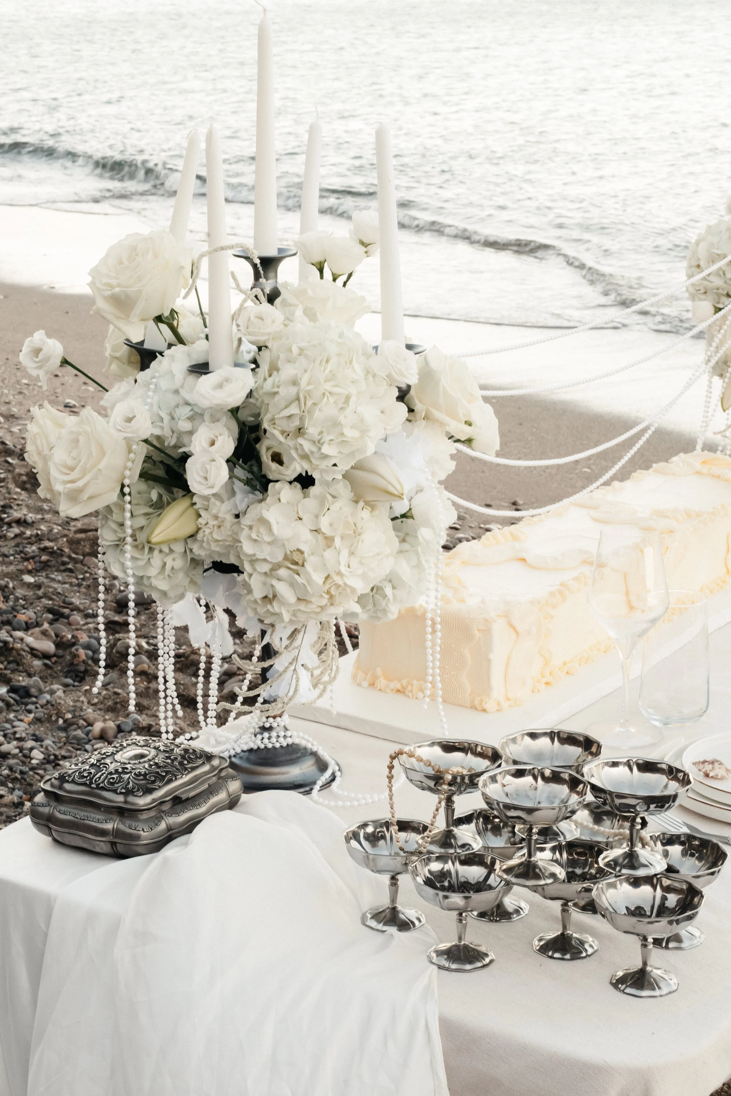 Décoration de mariage sur une plage avec un vase de fleurs blanches, chandeliers, assiettes en argent, gâteau blanc et verre à vin. Fond de mer avec petites vagues.