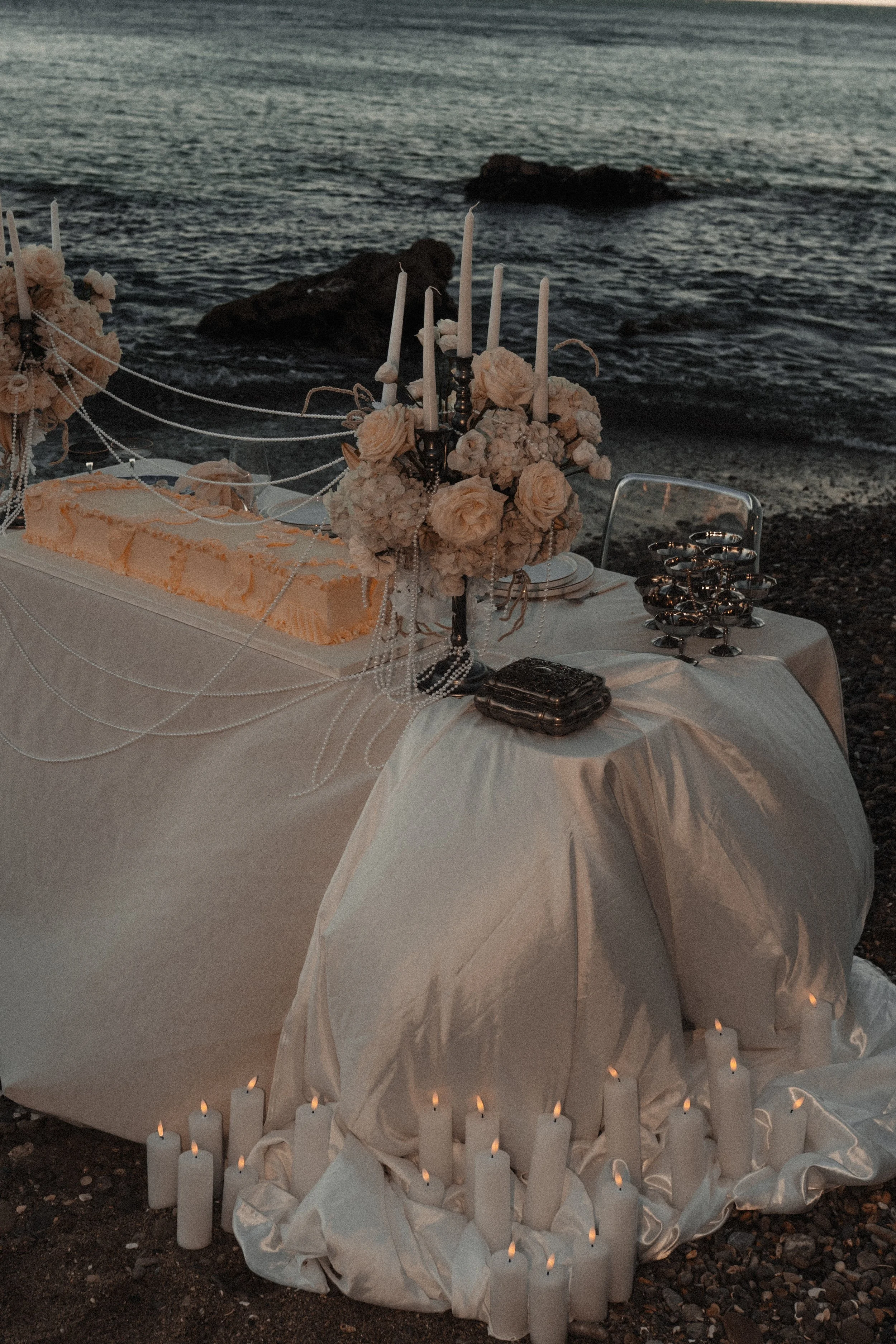 Table élégante dressée au bord de la mer avec des fleurs, bougies, candélabres, et un gâteau, pour une occasion spéciale ou une célébration.