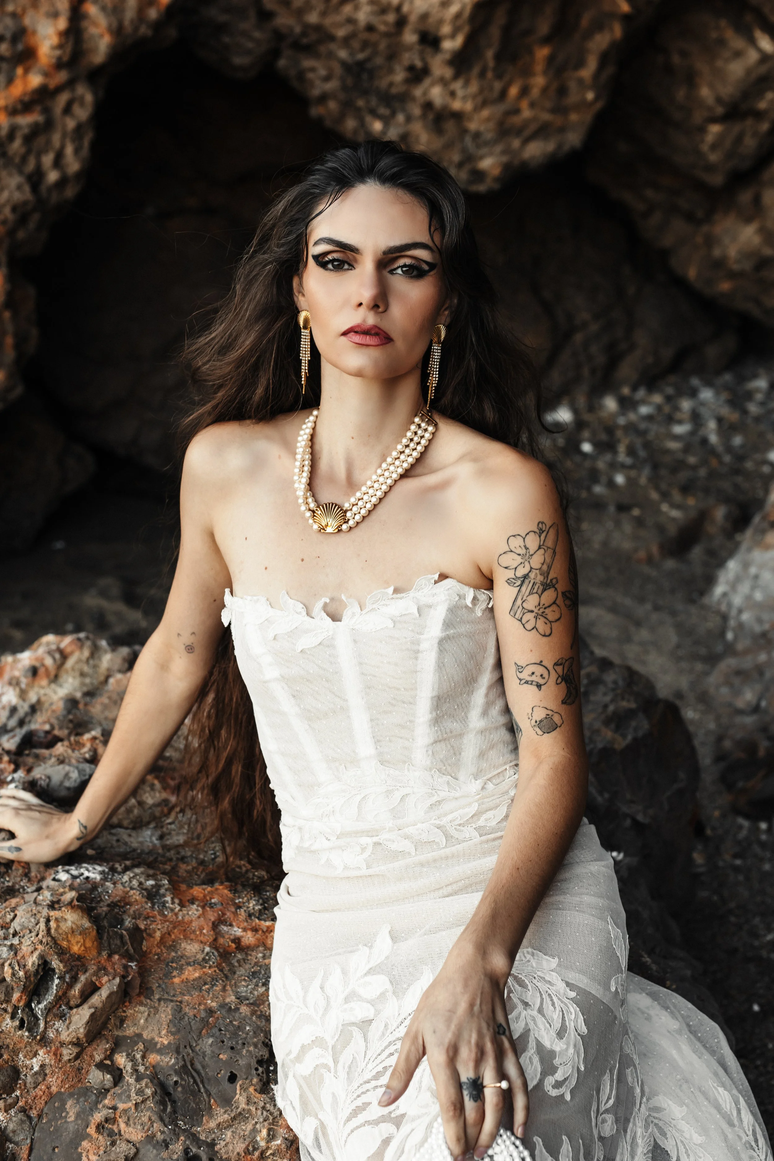 Femme portant une robe blanche en dentelle, avec des tatouages sur le bras, assise sur des rochers. Elle porte un collier de perles et des boucles d'oreilles longues, avec une atmosphère naturelle et sauvage.