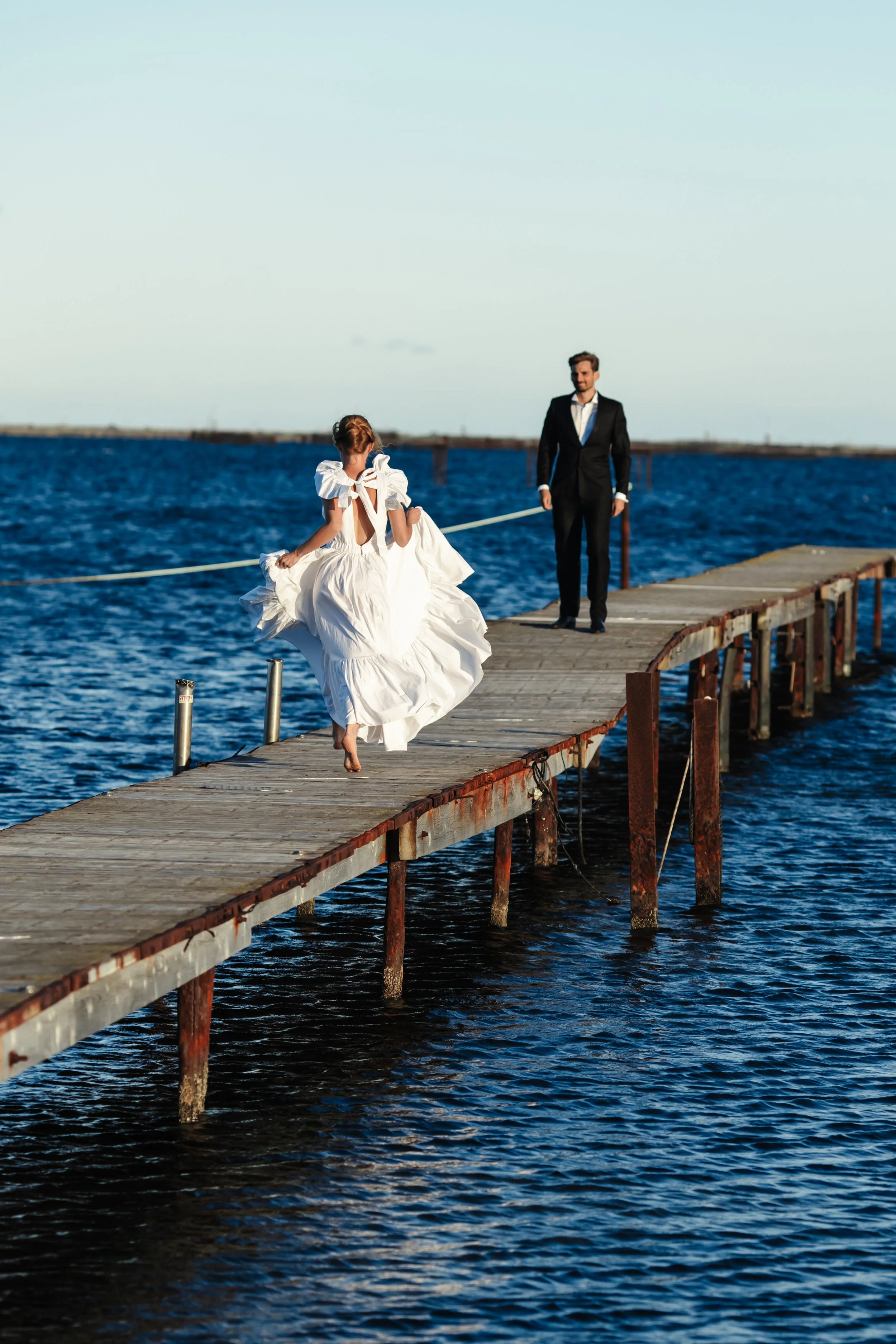Une femme en robe blanche décolle d'une jetée en bois lors d'une séance photo romantique, avec un homme en costume en arrière-plan, au bord de l'eau.