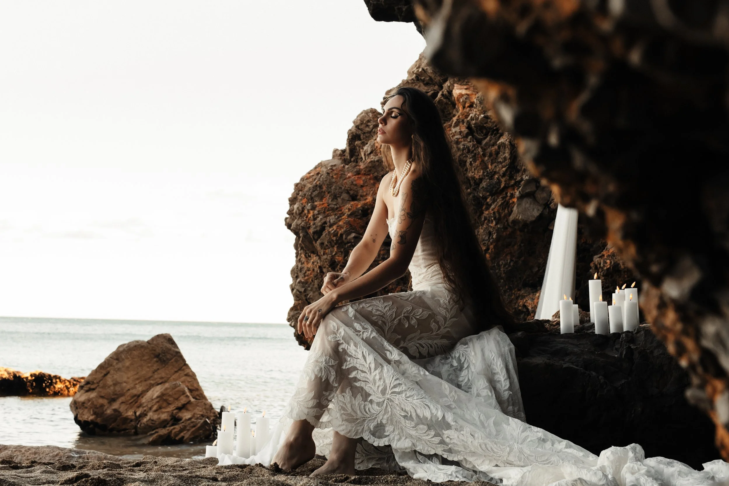 Femme en robe blanche assise sur le sable d'une plage rocheuse, entourée de bougies allumées, avec la mer en arrière-plan.