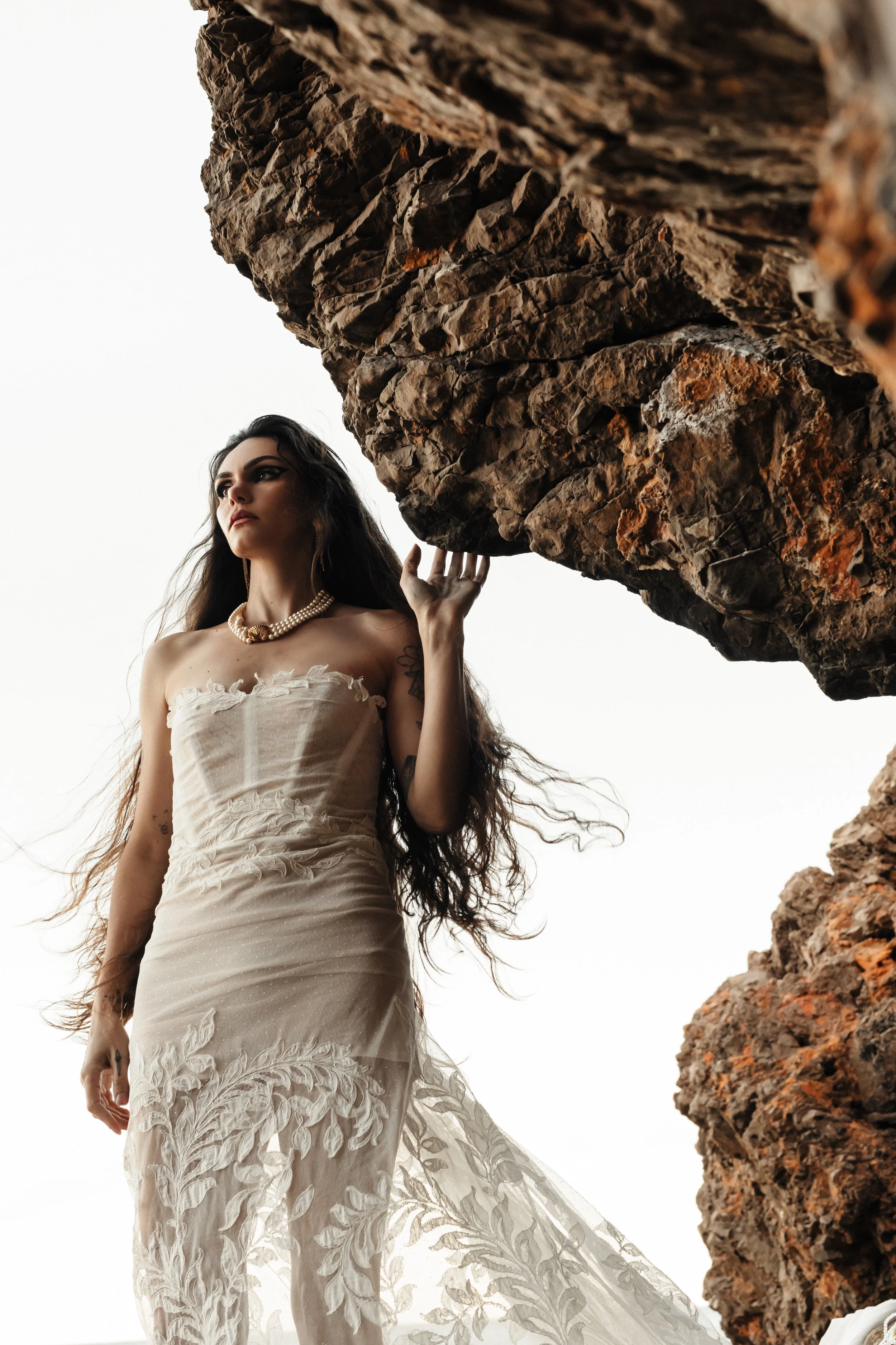 Une femme portant une robe en tissu léger et des bijoux, debout près d'une formation rocheuse, regardant au loin.