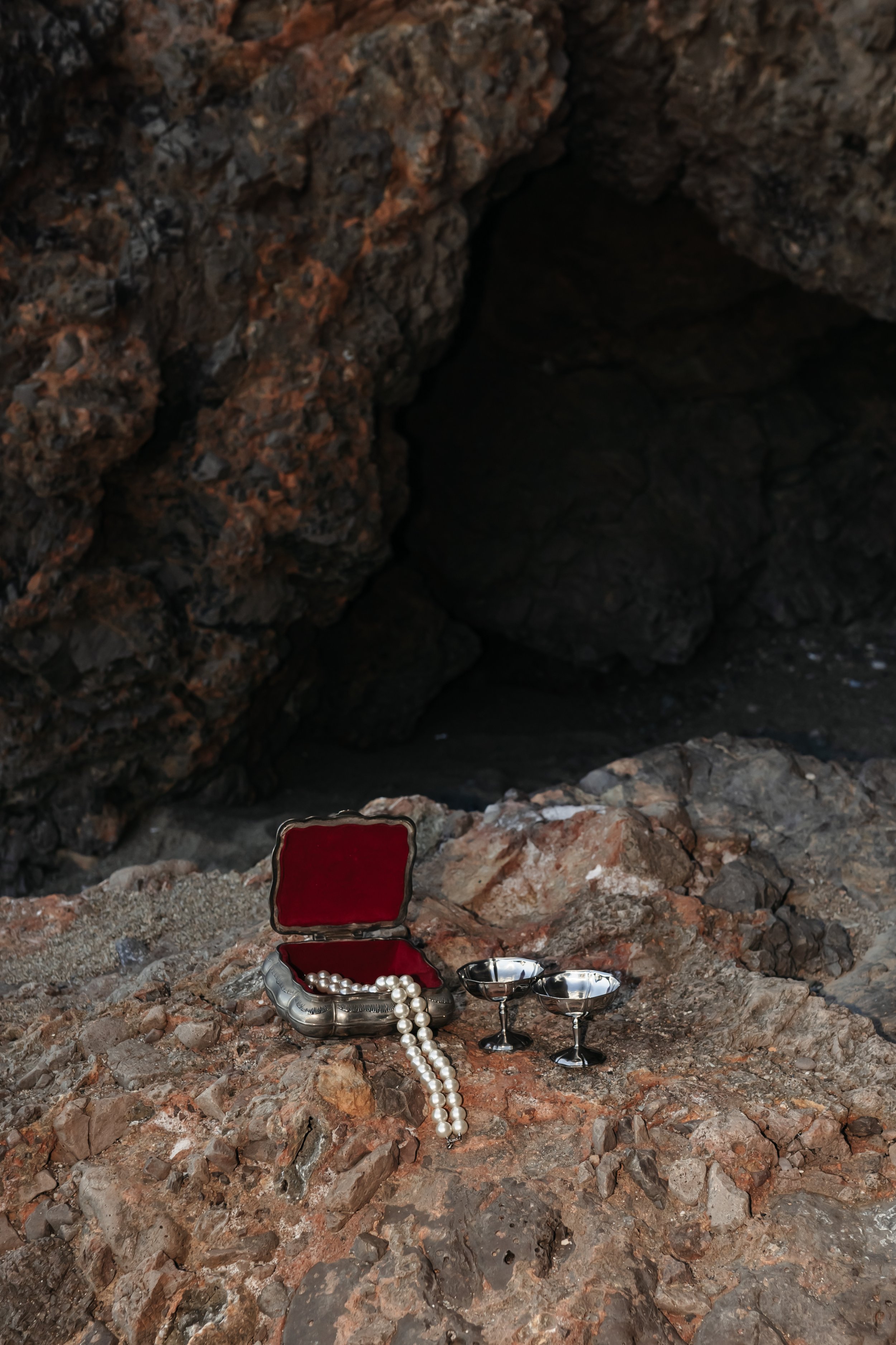Une boîte en forme de coquillage en argent avec un collier de perles, deux verres à vin en argent, tous placés sur des rochers au bord d'une grotte ou d'un trou dans la roche.