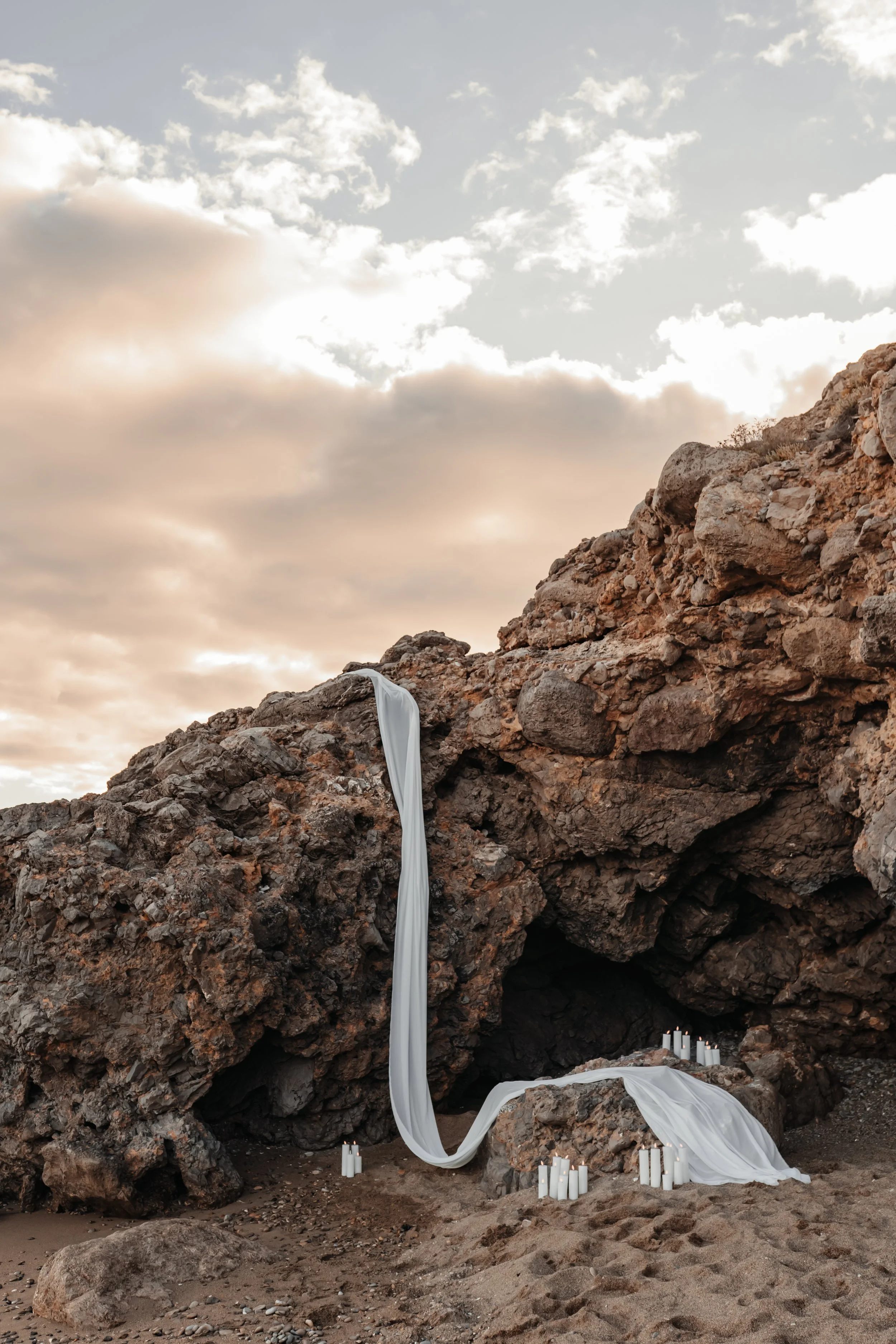 Décoration nuptiale sur la plage avec un rocher, d'une étole blanche suspendue entre deux rochers, entourée de bougies