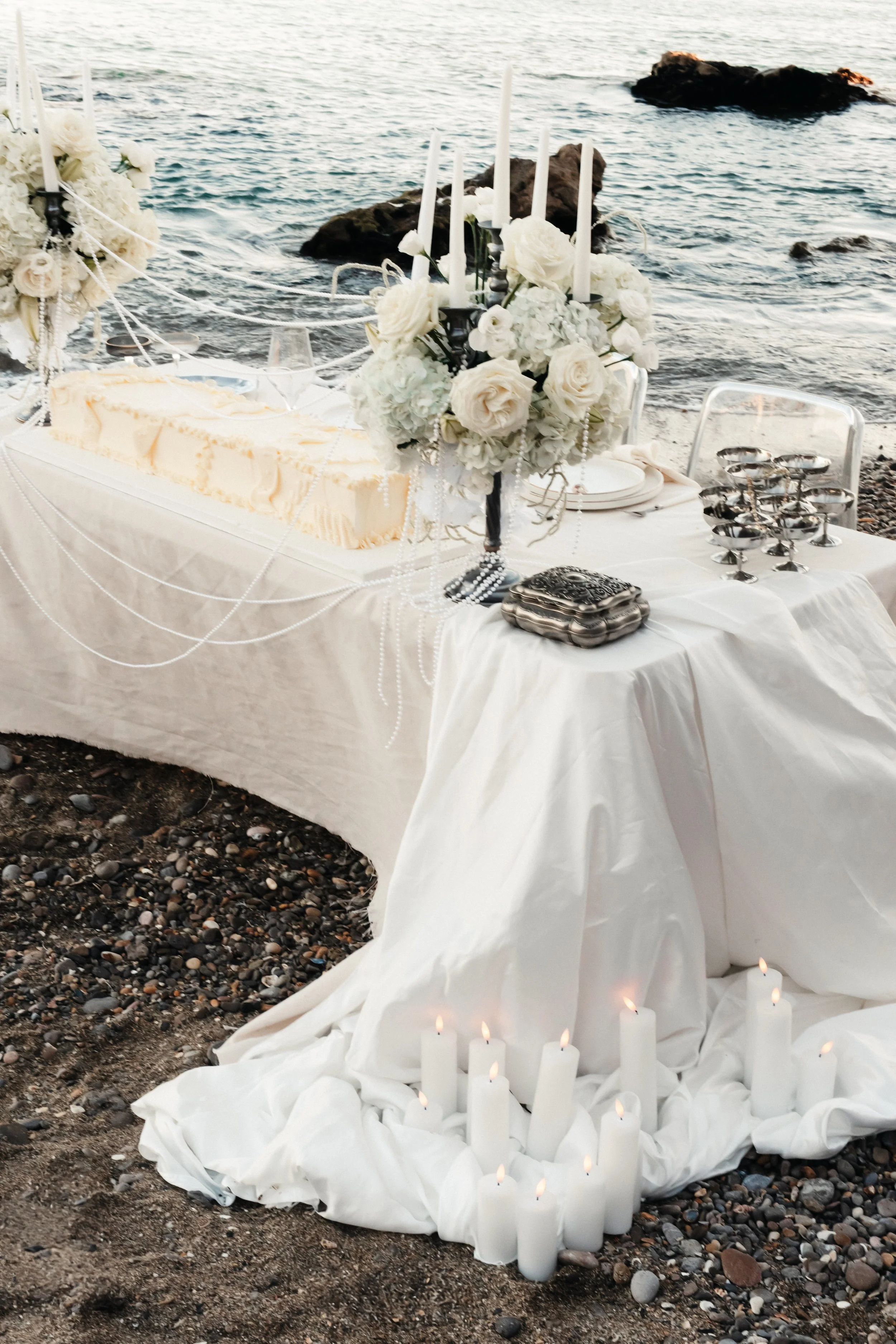 Table décorée avec des fleurs blanches, des bougies et de la vaisselle, située sur la plage près de la mer.