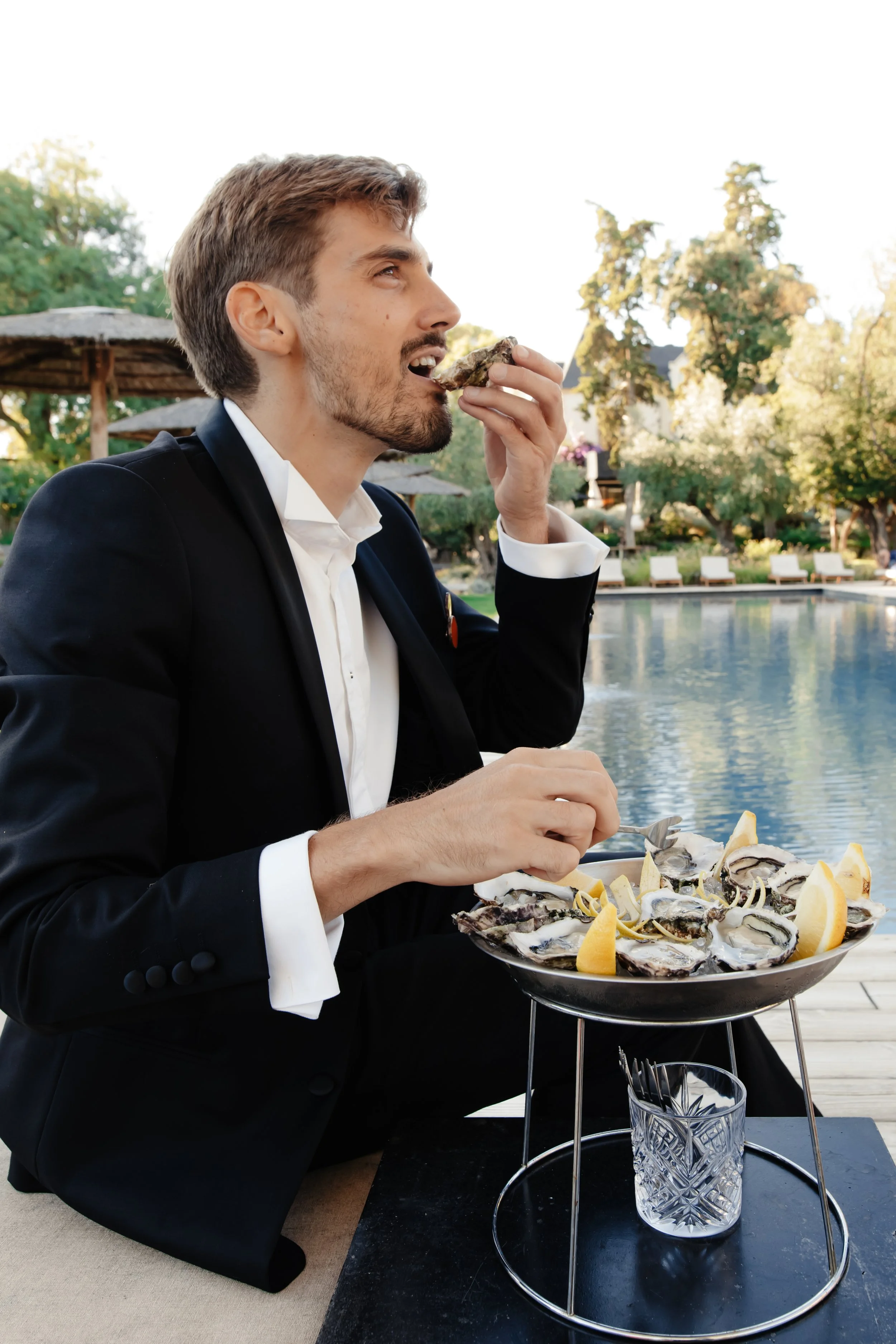 Un homme habillé en costume noir mange des huîtres avec du citron à l'extérieur près d'une piscine