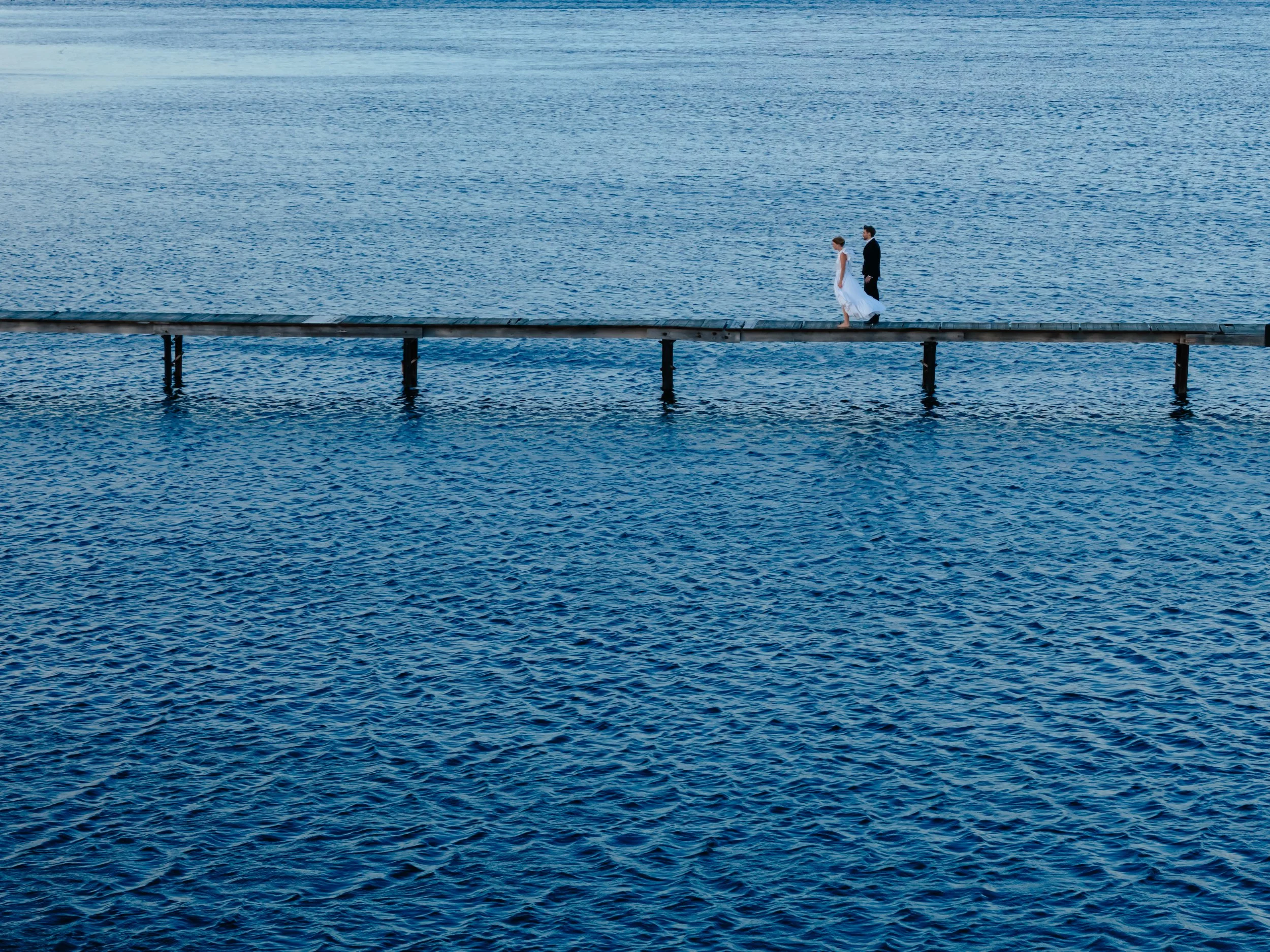 Un couple marié marche sur une jetée en bois au-dessus d'une mer calme, vue panoramique.