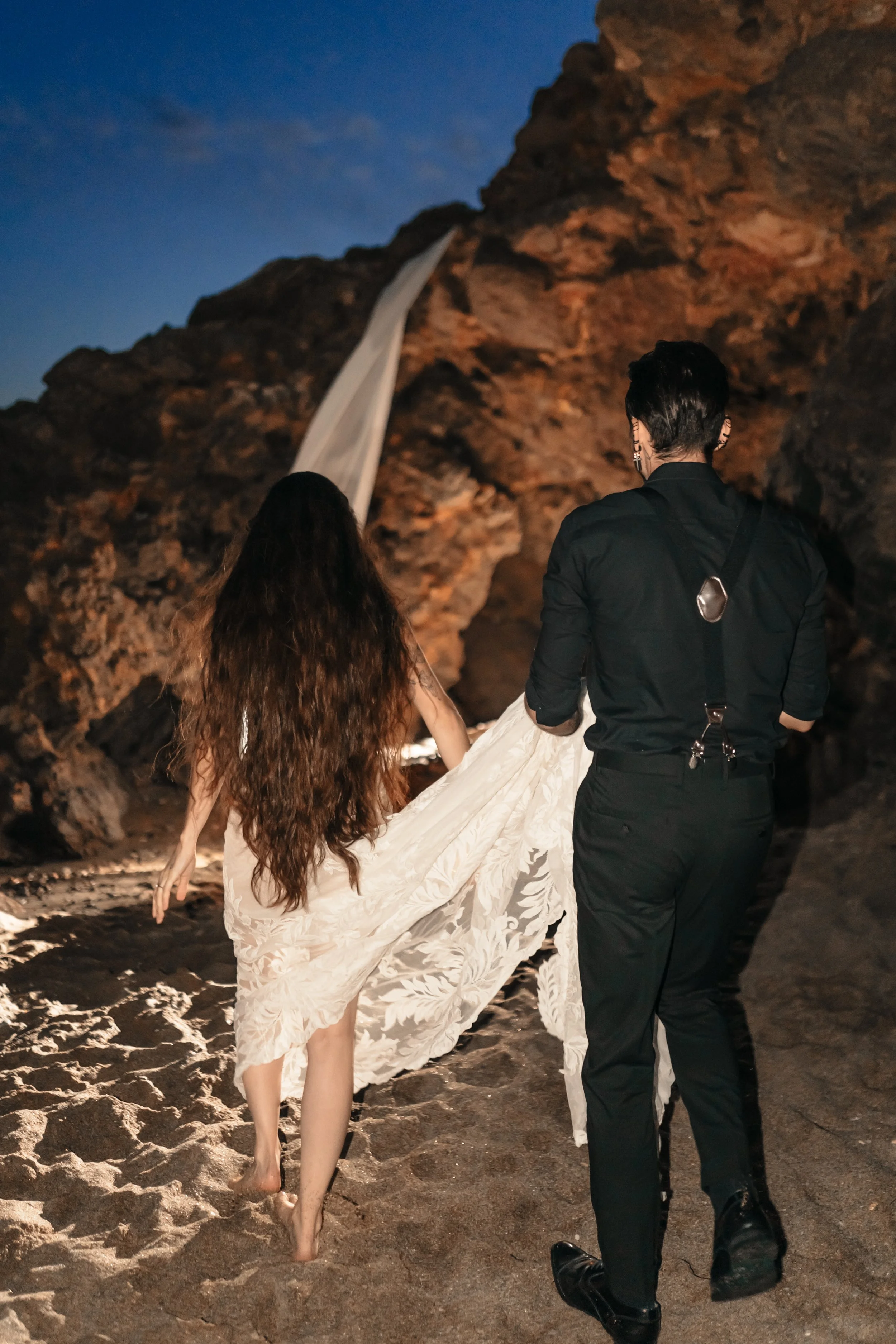 Deux personnes, dont une en robe blanche, marchent pieds nus sur une plage rocheuse au crépuscule.