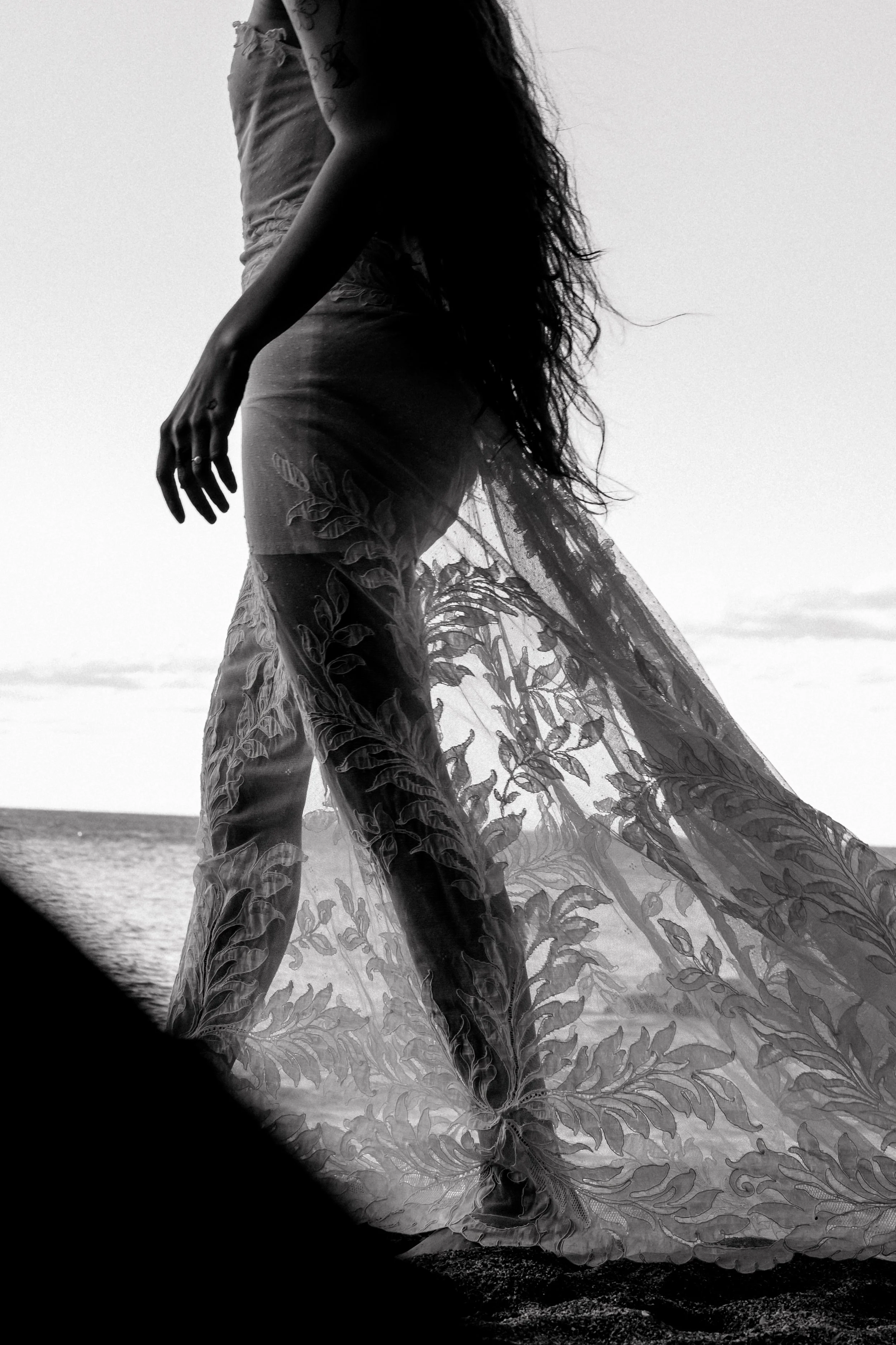 Une femme portant une robe longue en tissu transparent avec des motifs floraux, debout près de la mer au coucher du soleil.